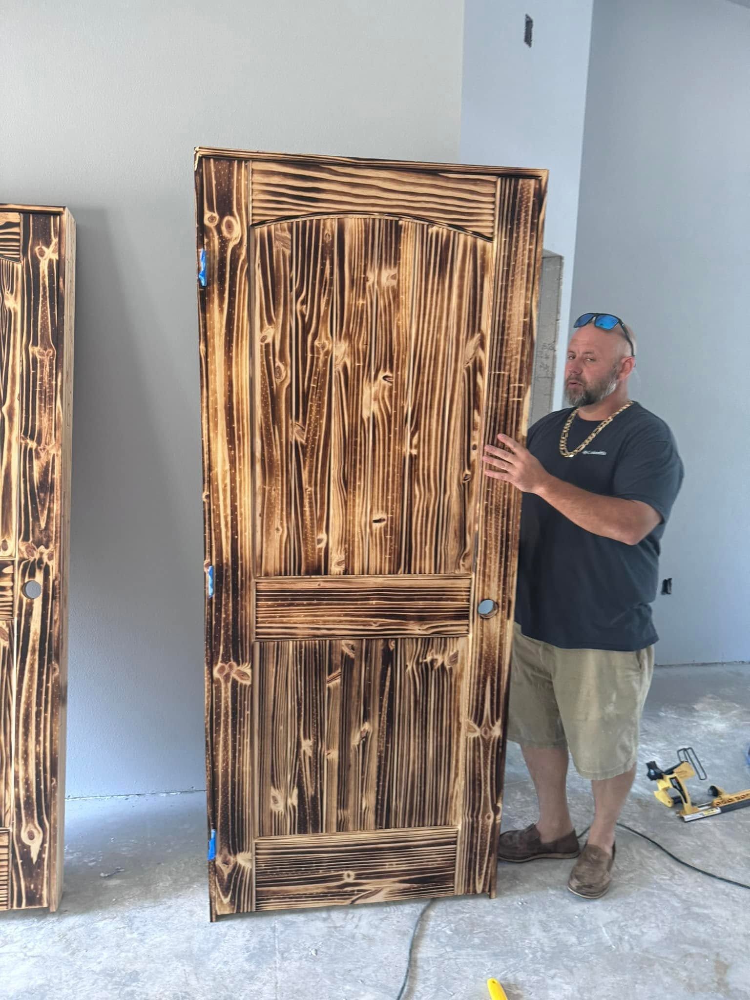 Man standing beside a large rustic wooden door with dark stain in a workshop-like room