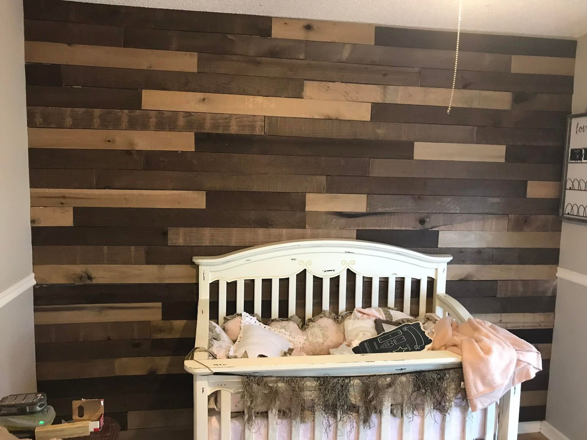Baby sleeping in a white crib against a dark wood plank wall, with a pink blanket draped over the side.
