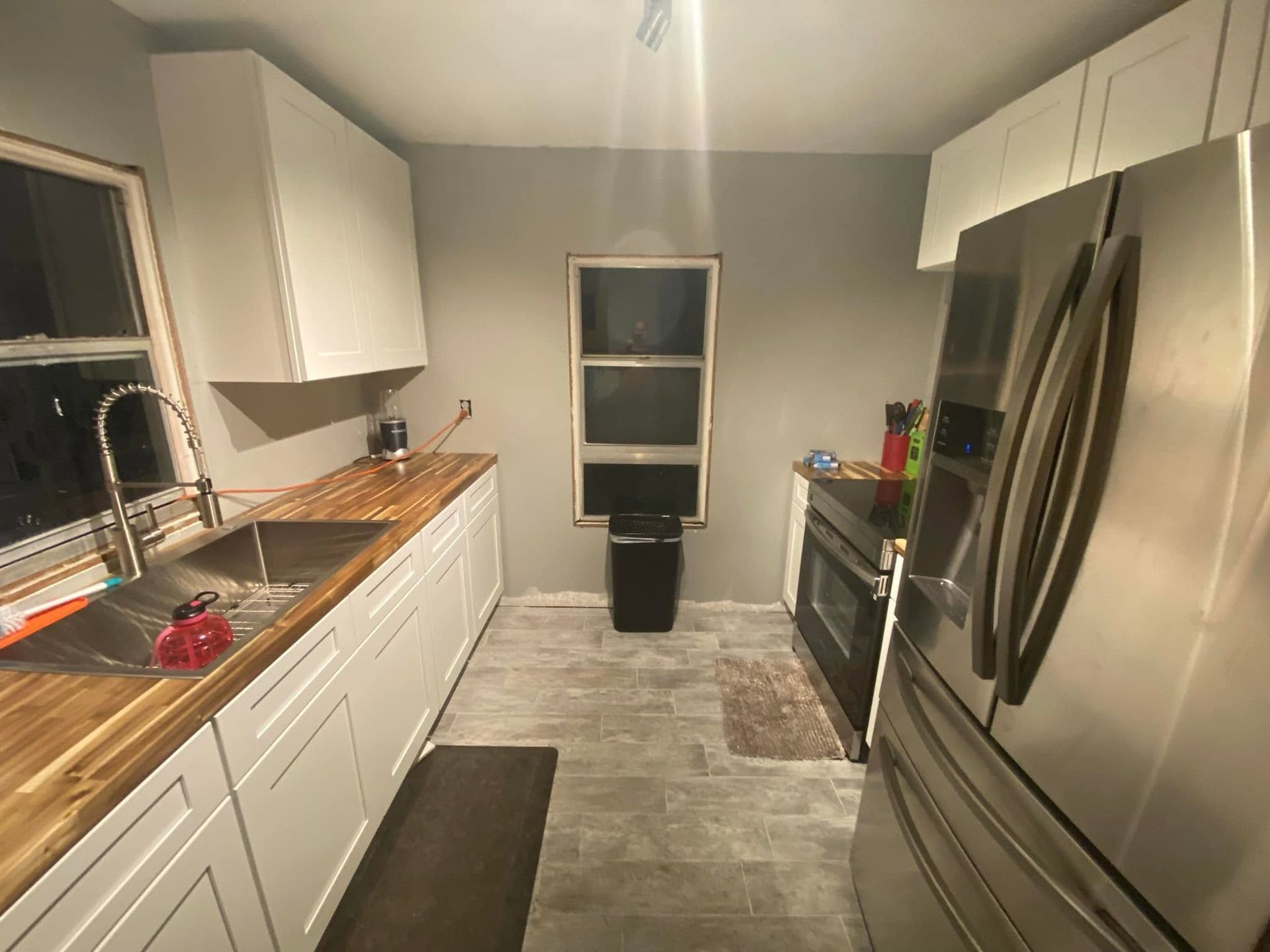 Narrow galley kitchen with white cabinets, stainless steel fridge, and a window over the sink.