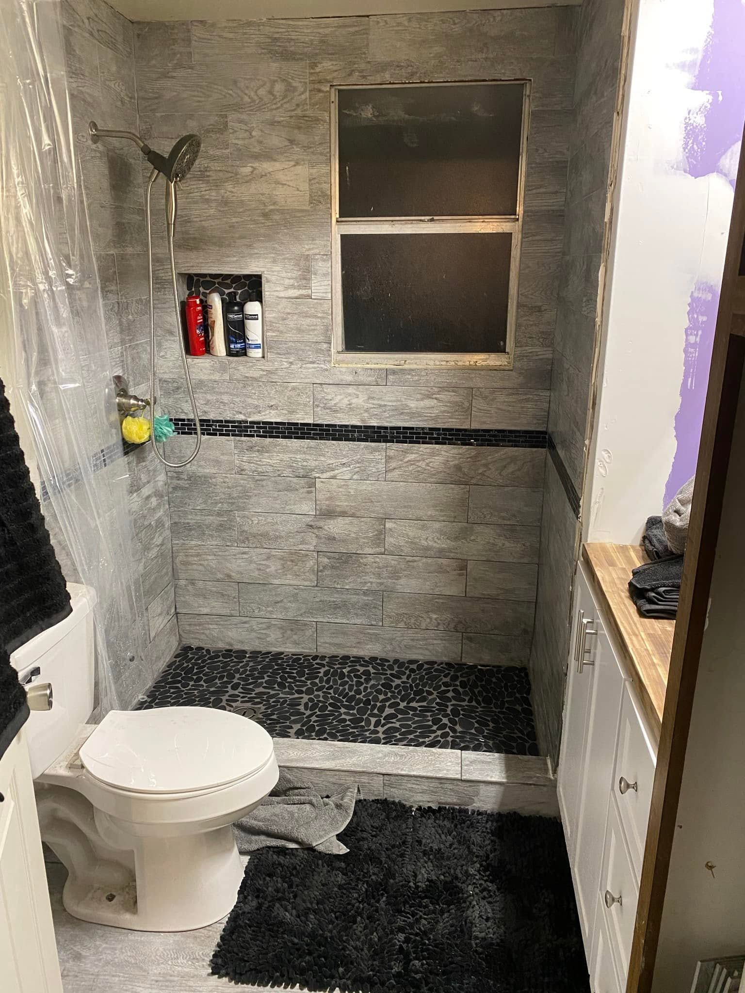 Small tiled bathroom with a toilet, shower alcove, window, and gray stone-pattern walls.