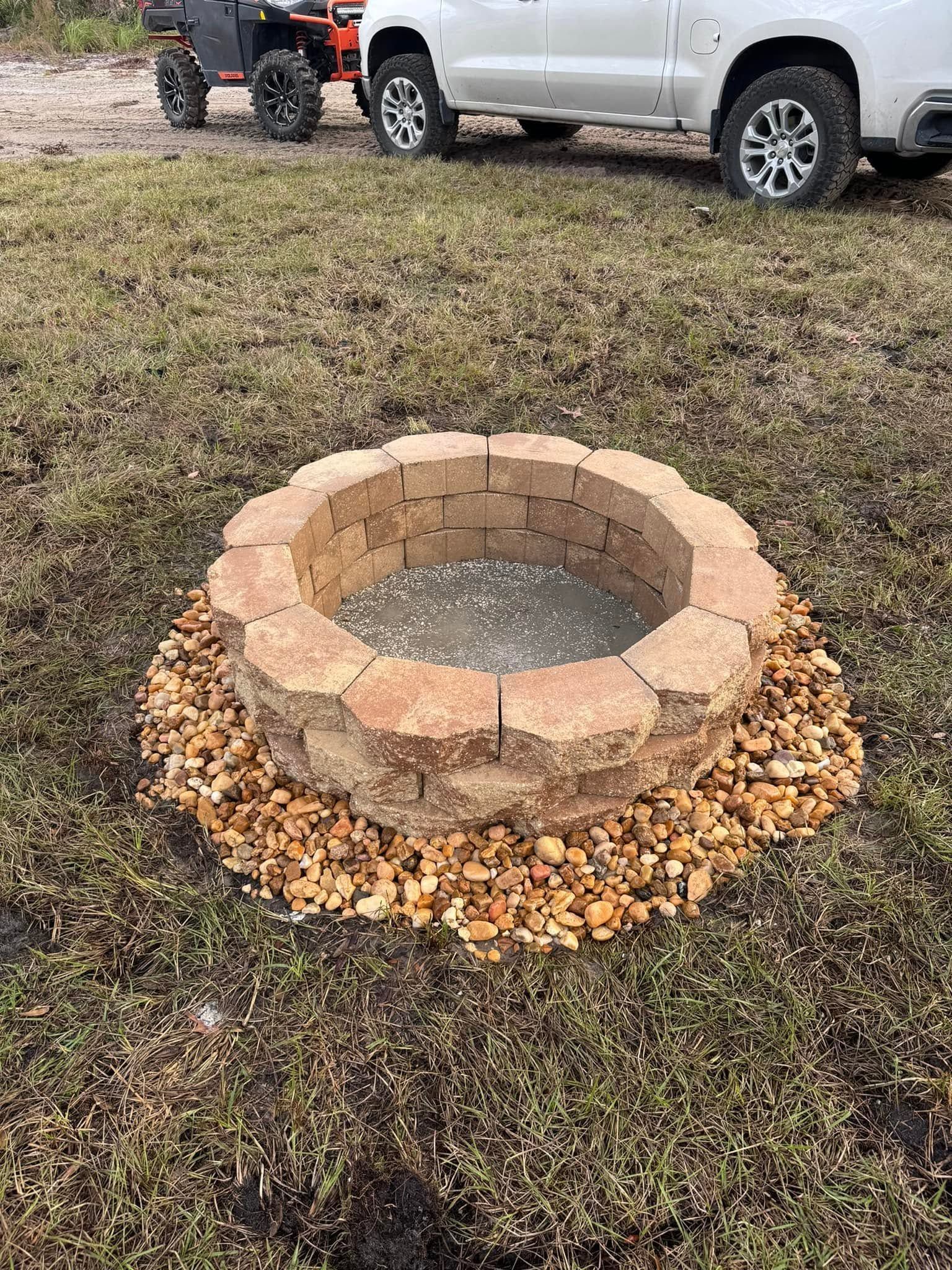 Small stone fire pit ringed with gravel on muddy grass, with a white pickup truck in the background
