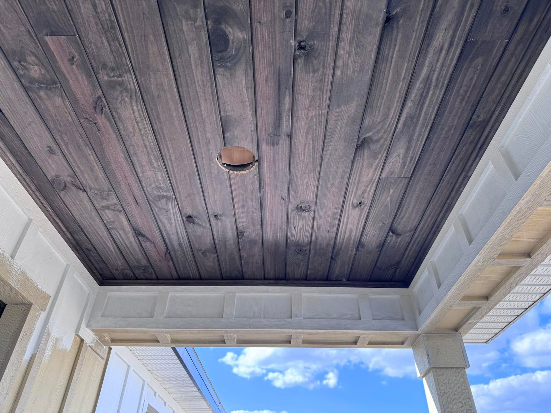 Dark wood porch ceiling with a small light fixture above white trim and a glimpse of blue sky.