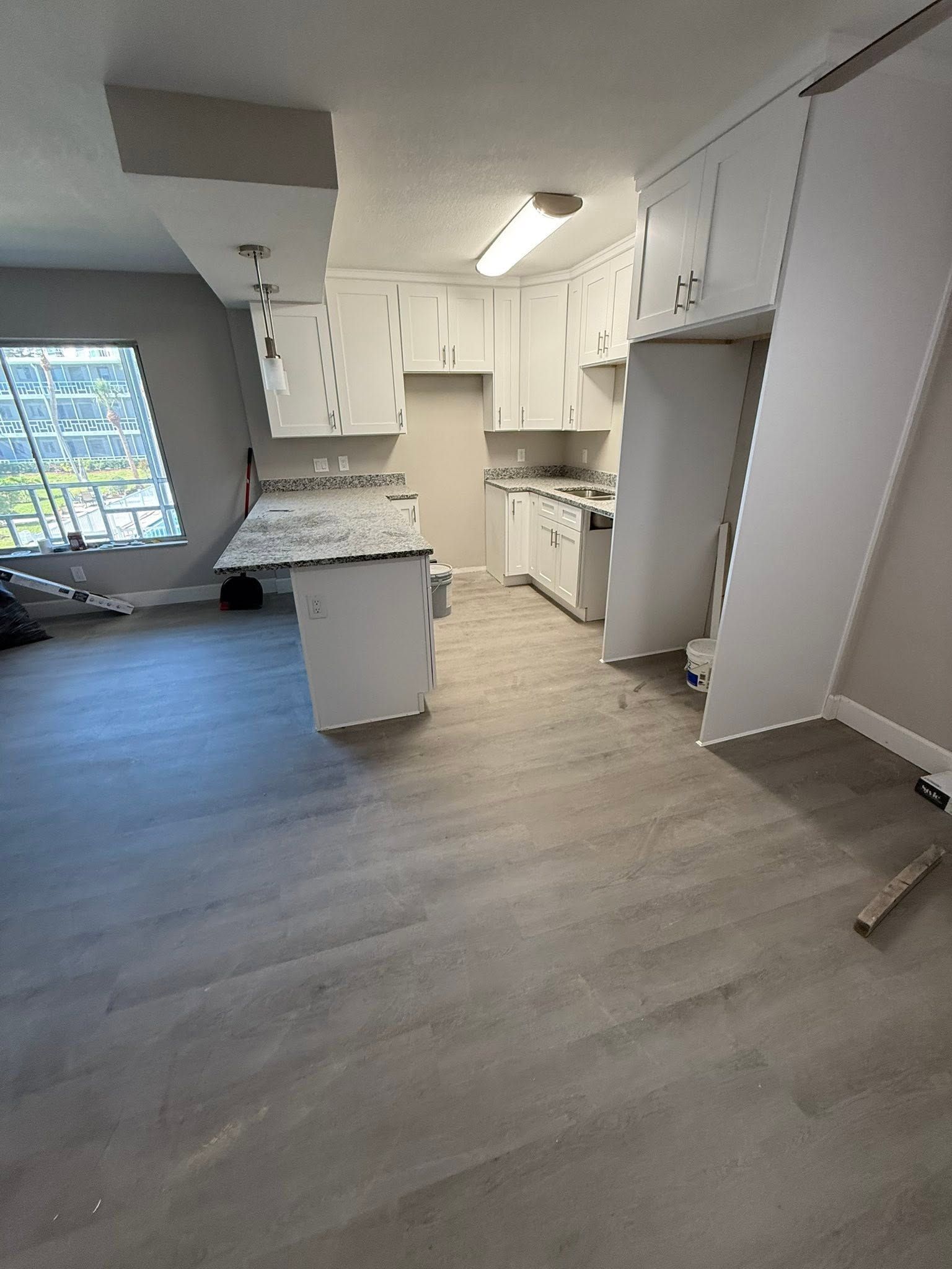 Empty open-plan kitchen with white cabinets, gray flooring, and a large window.