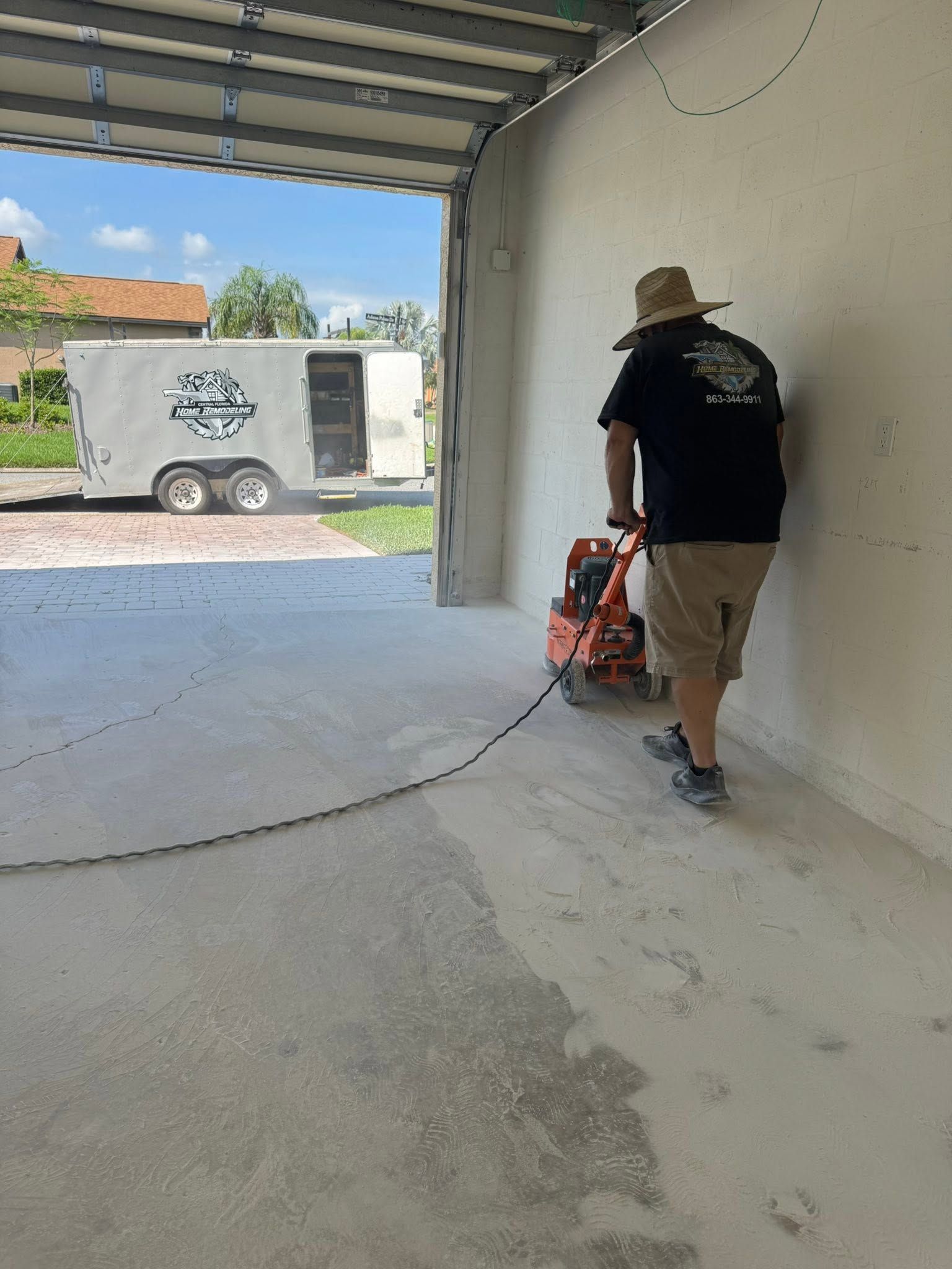 Worker using a red floor buffer in an empty garage bay with an open door to a truck outside