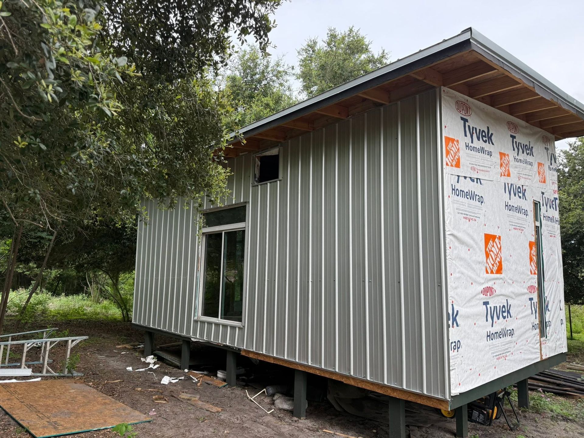 Small gray metal cabin under construction in a wooded area, with Tyvek wrap on the end wall.