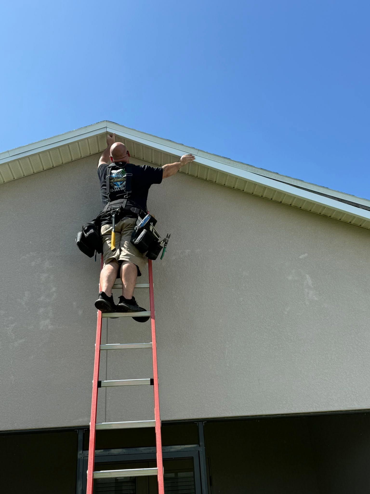 Worker on a ladder repairing the roof trim of a house under a clear blue sky