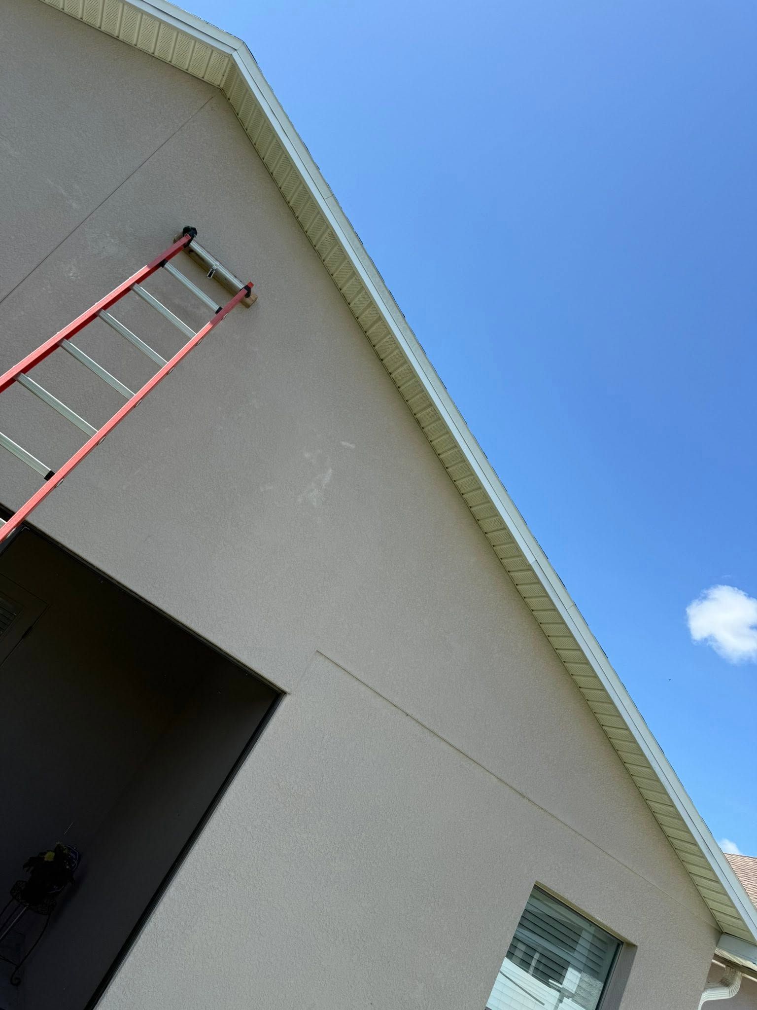 Red ladder leaning against a gray stucco house under a blue sky with a small cloud