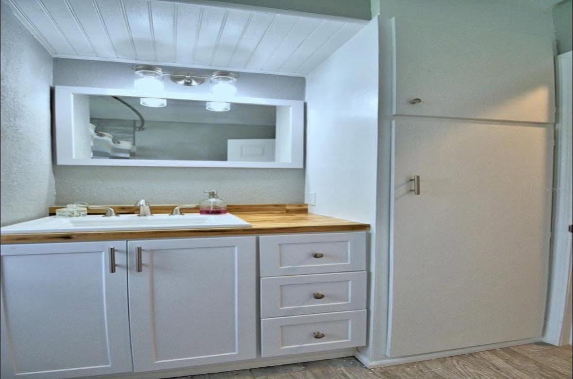 Small white bathroom vanity with sink, mirror, wood countertop, and adjacent cabinet wall