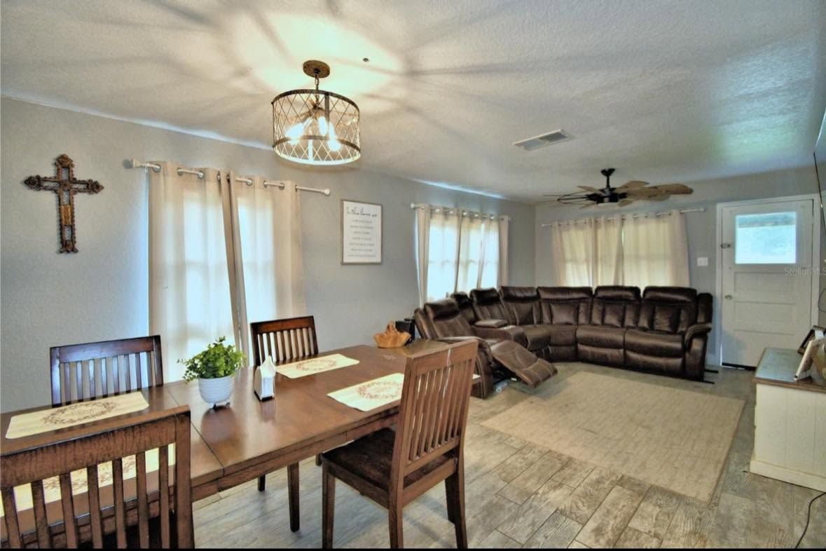 Bright living and dining room with wooden table, sectional sofa, wall cross, and large windows.
