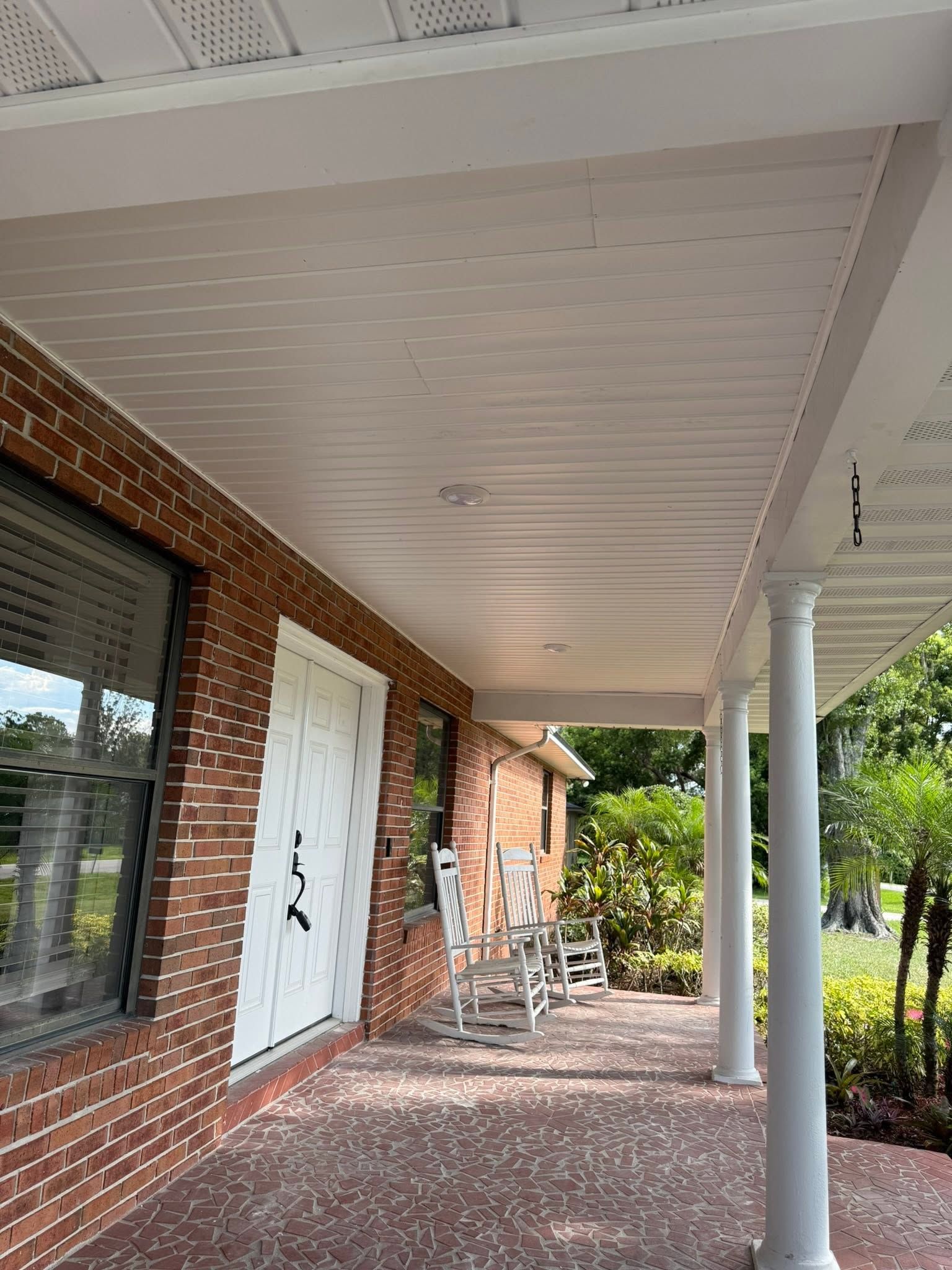 Brick porch with white ceiling, white door, rocking chairs, and a white column beside greenery.