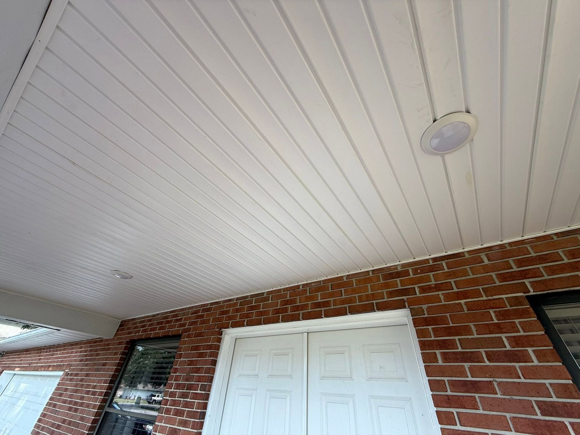 Covered porch with white beadboard ceiling, brick wall, and double white doors.