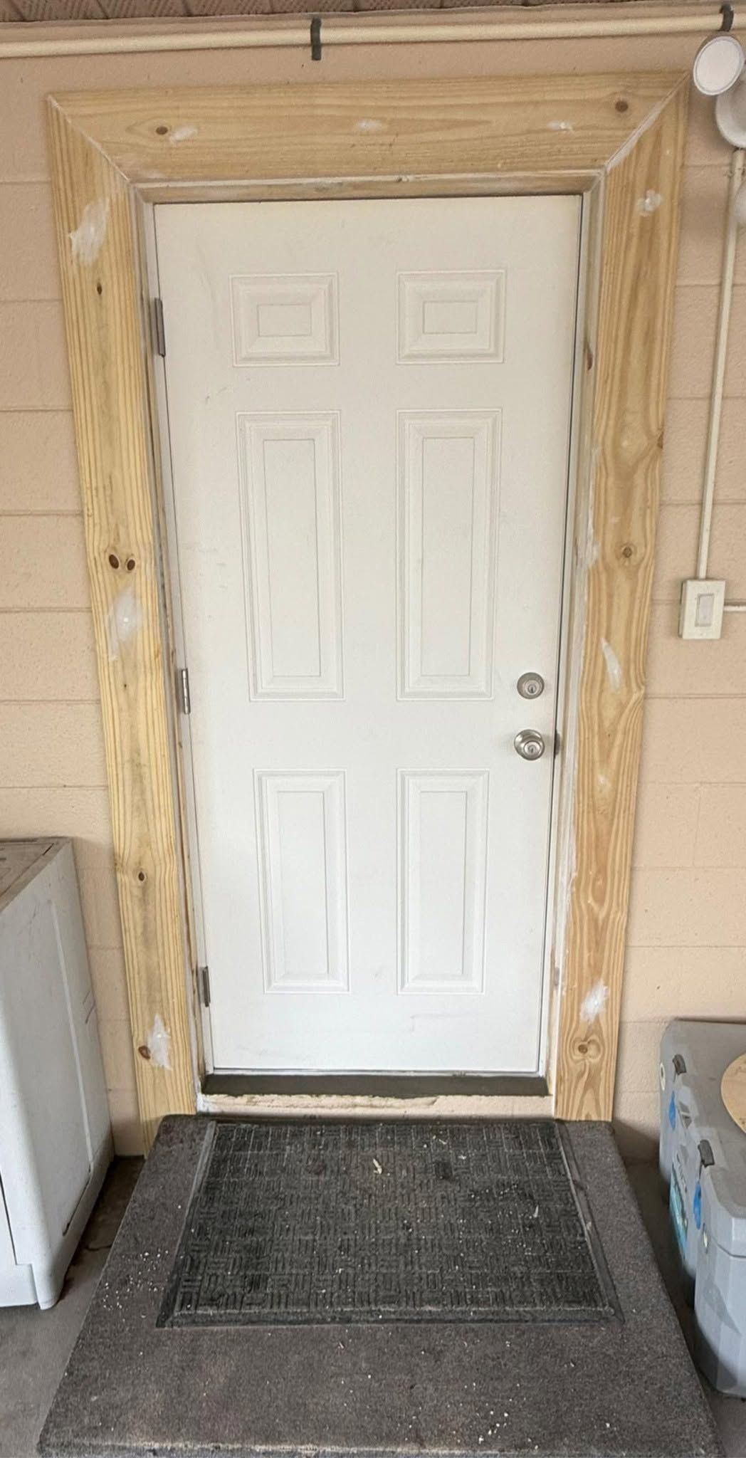 White exterior door with wooden framing, a doormat, and a wall-mounted switch beside it
