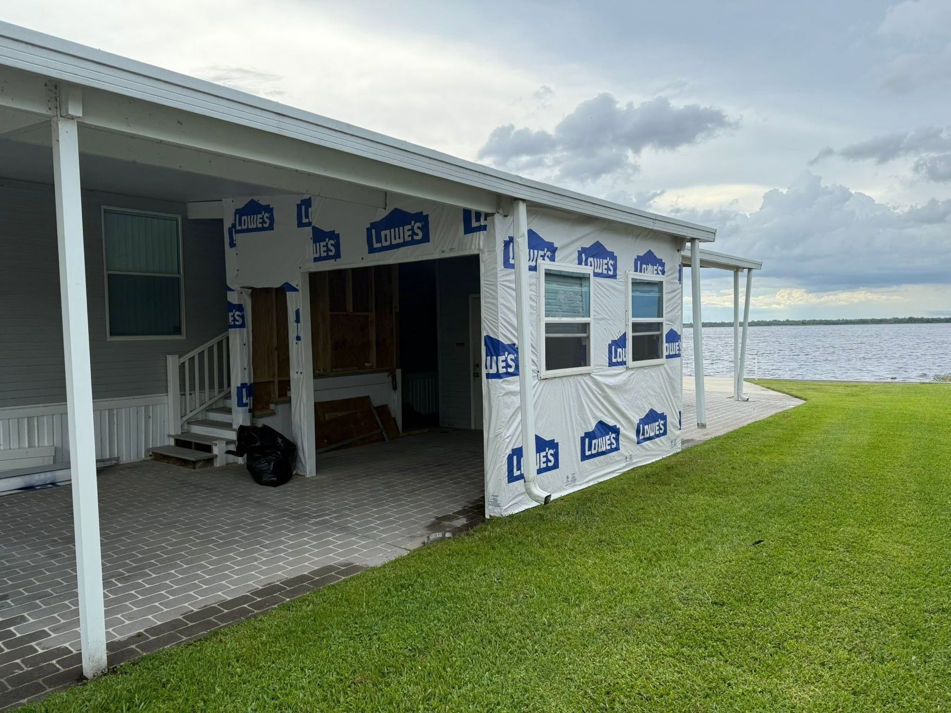 Porch of a lakeside cabin under construction, with white siding, blue trim, and grassy shoreline nearby.