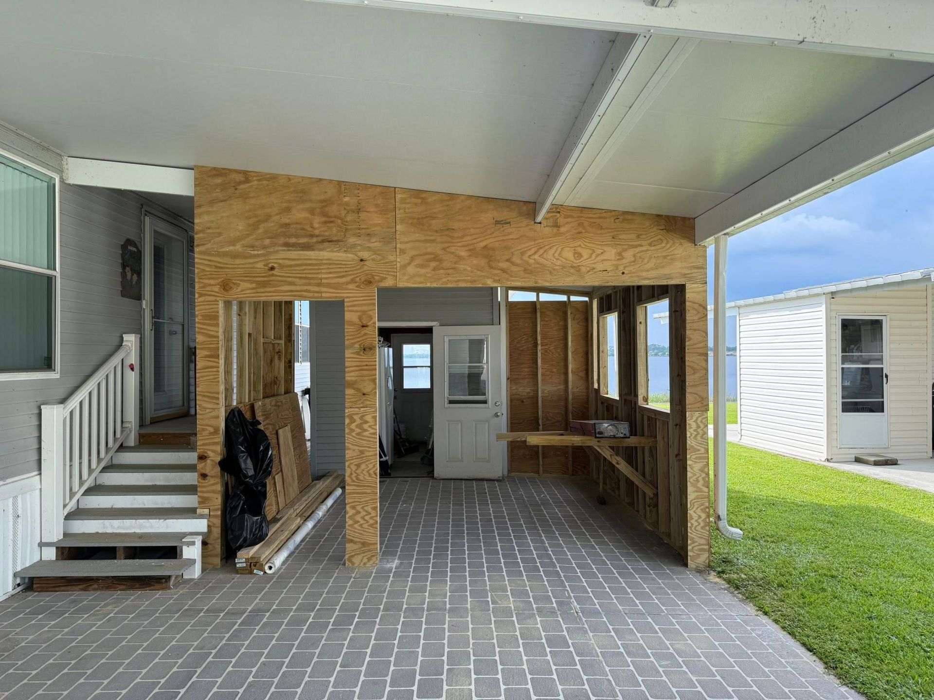 Covered outdoor patio with unfinished wood walls, white stairs, and a paved floor beside a grassy yard.