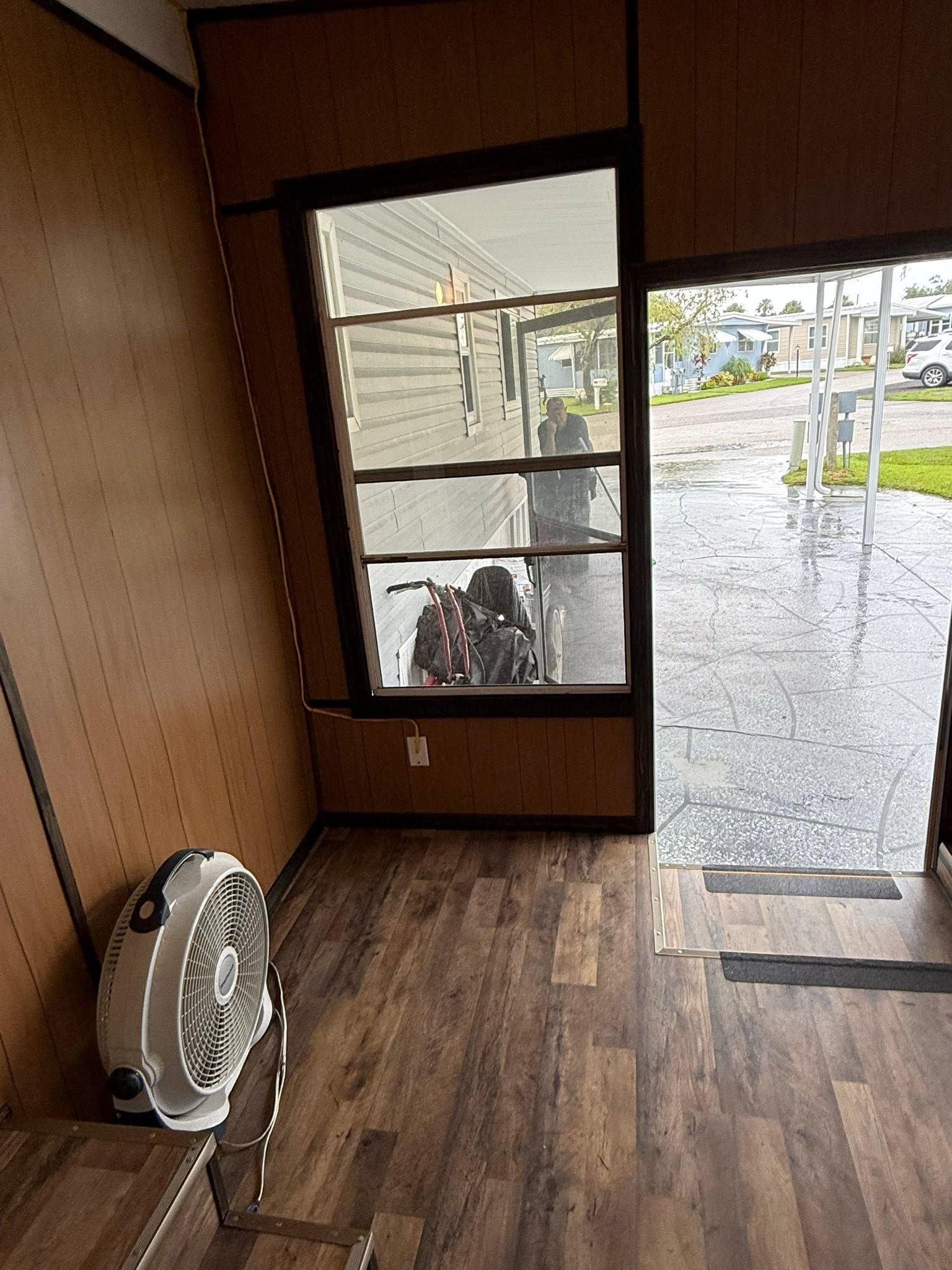 Empty room with wood floors, fan on the left, and an open glass door to a rainy street.