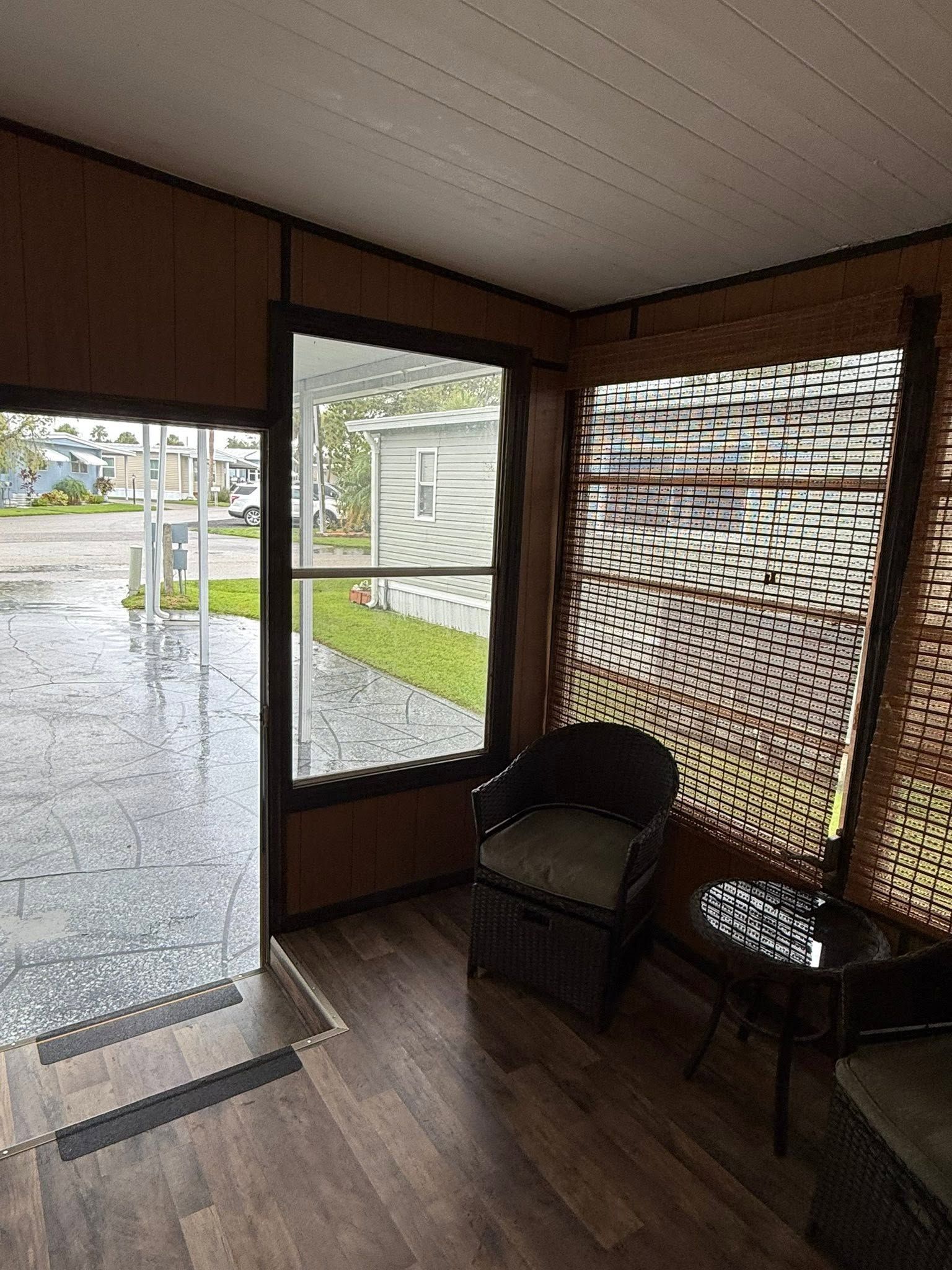 Small covered porch with two chairs, a woven screen, and an open door looking out to a gravel yard.