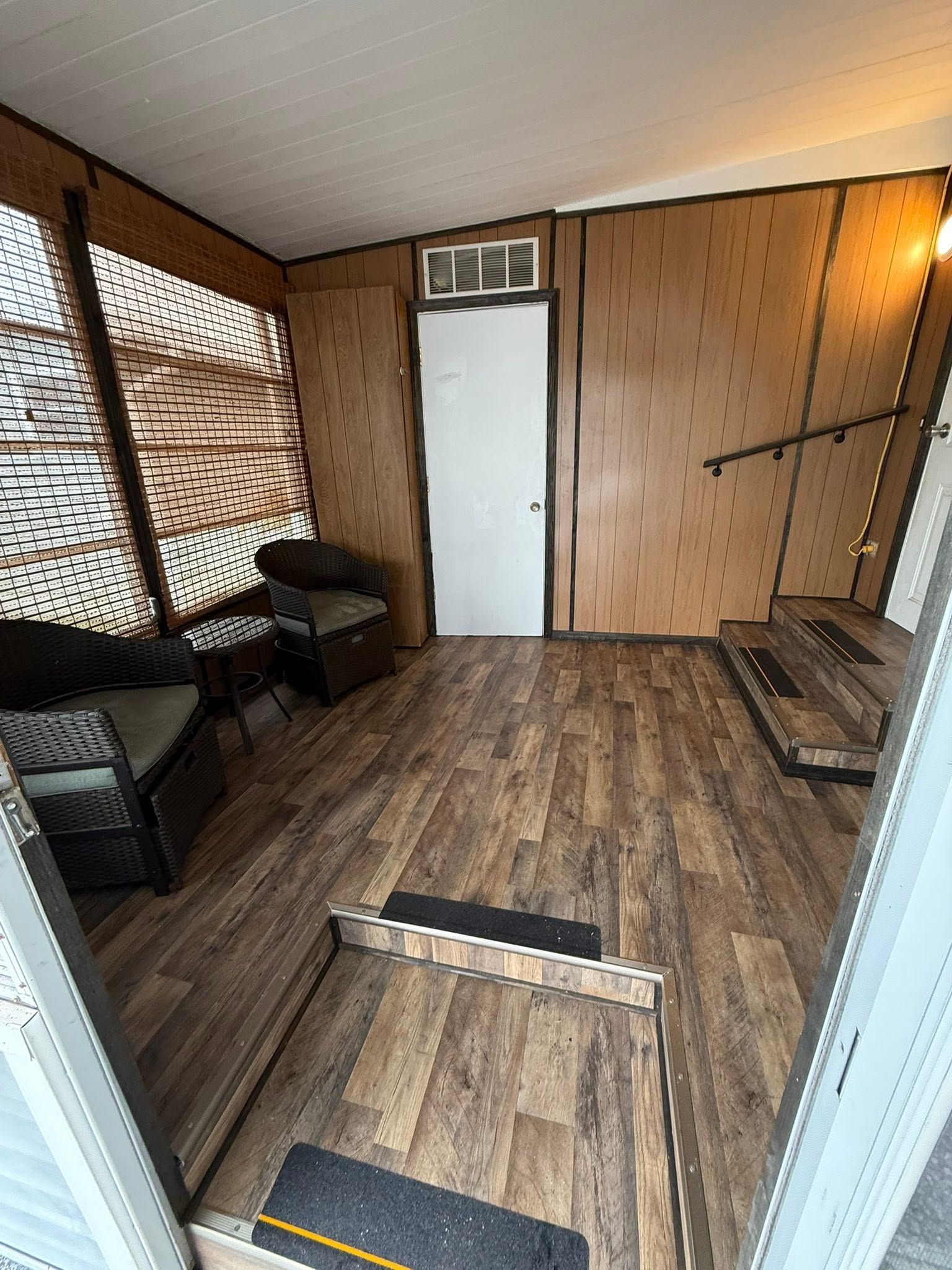 Small wood-paneled room with a white door, chairs, a bench, and a damaged floorboard.