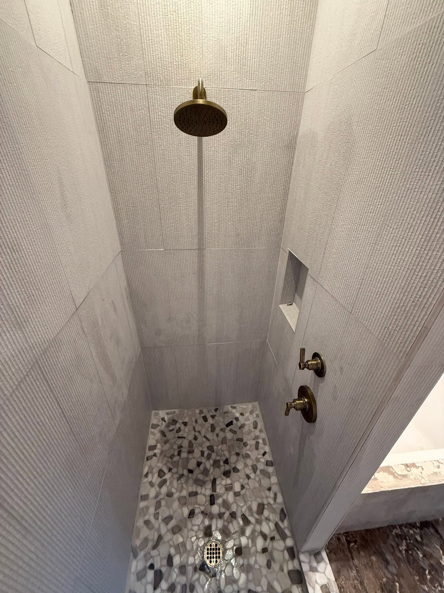 Small tiled shower stall with patterned wall tiles, brass shower head, and mosaic floor.