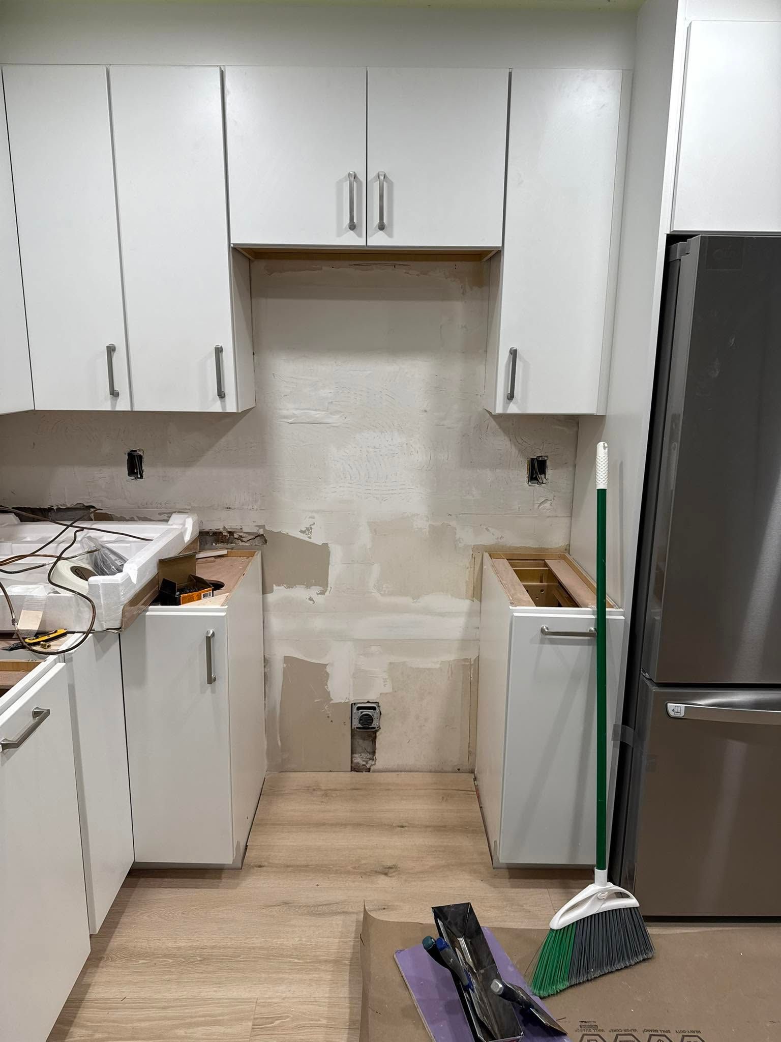 Narrow kitchen under renovation with white cabinets, exposed backsplash, tools, and a refrigerator on the right.