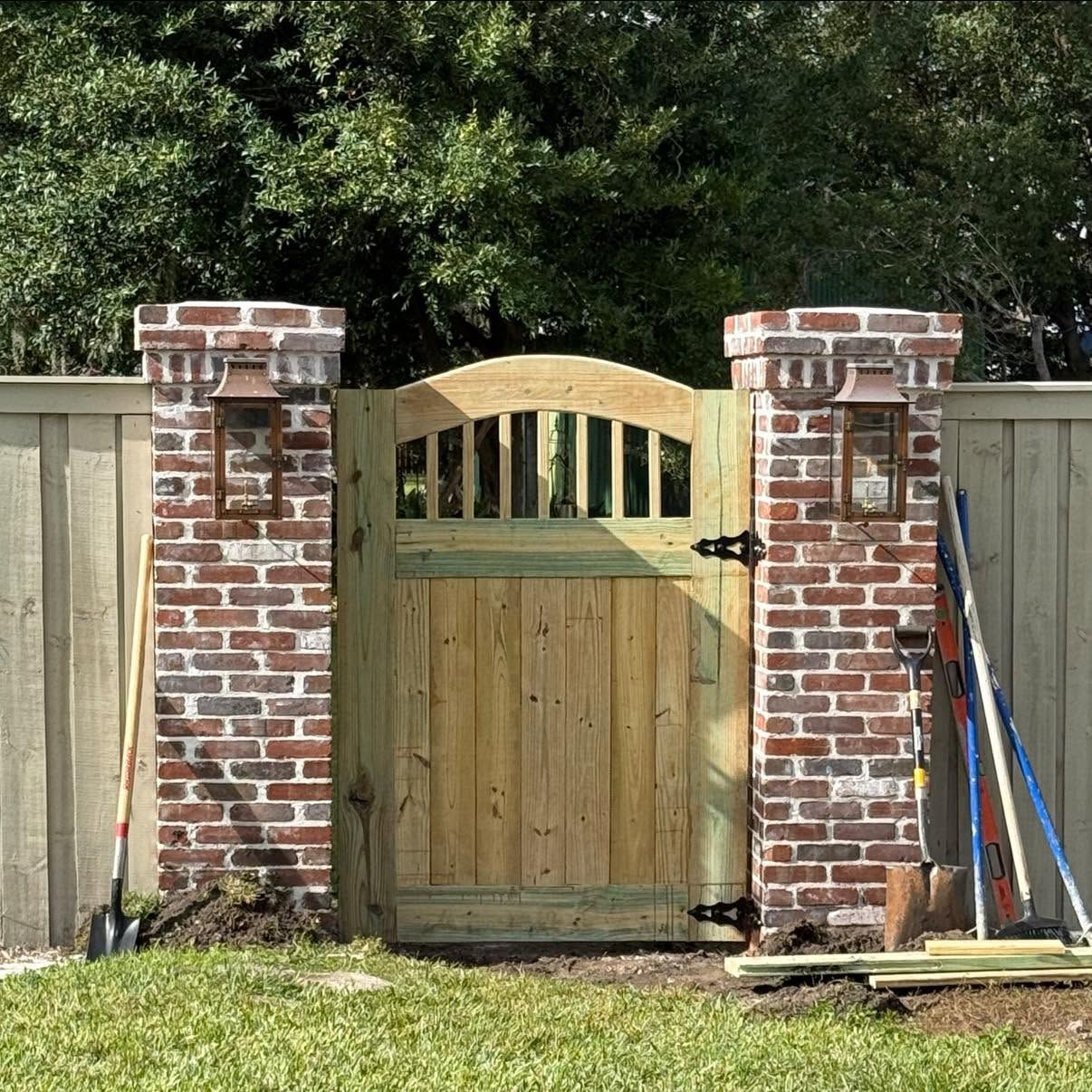 Wooden gate between two brick pillars in a fenced backyard, with trees and lawn behind it