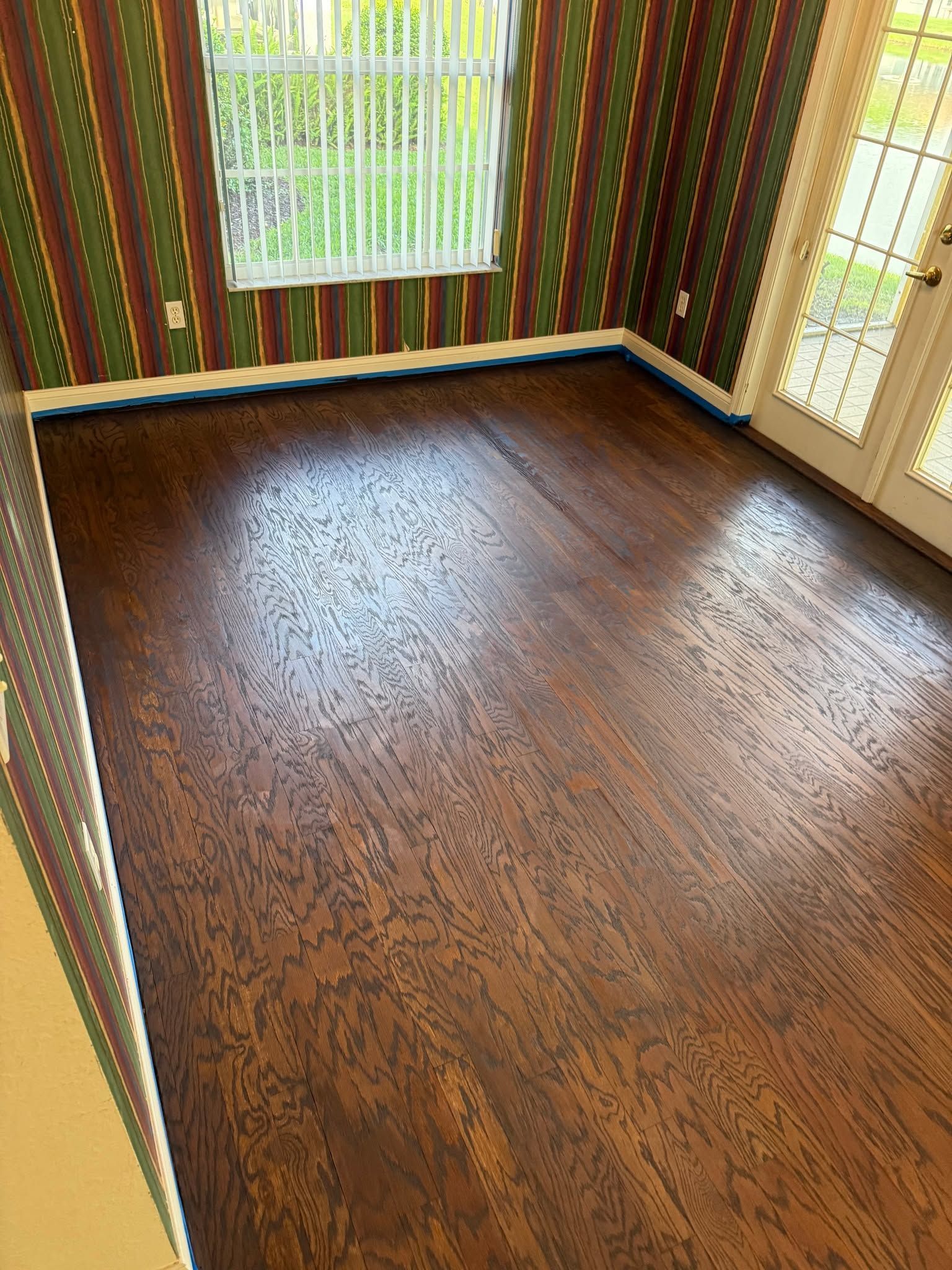 Empty hardwood room with striped wallpaper and two windows, one with a white blind.