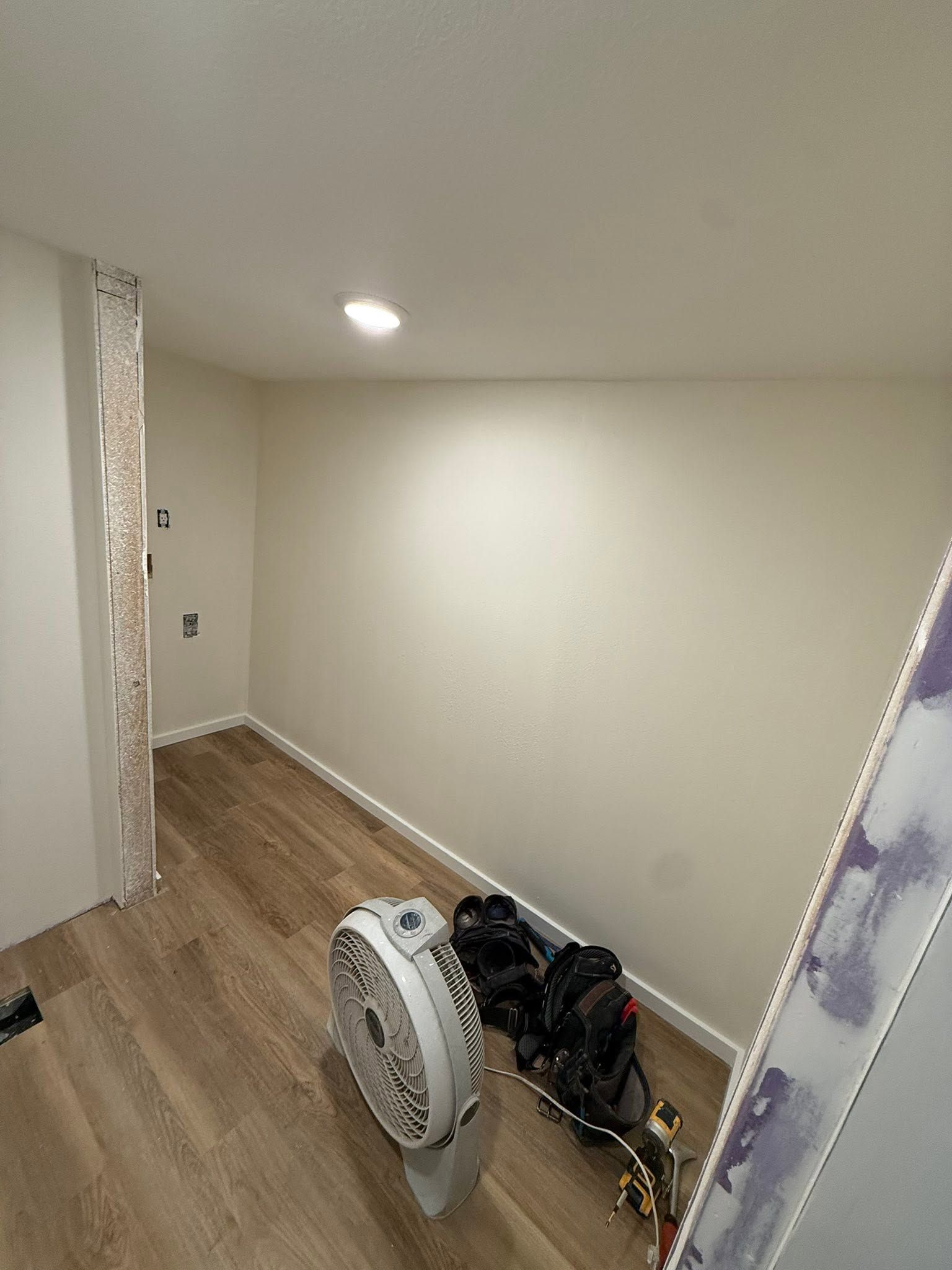 Small empty room with wood floor, cream walls, and a portable fan and equipment on the floor