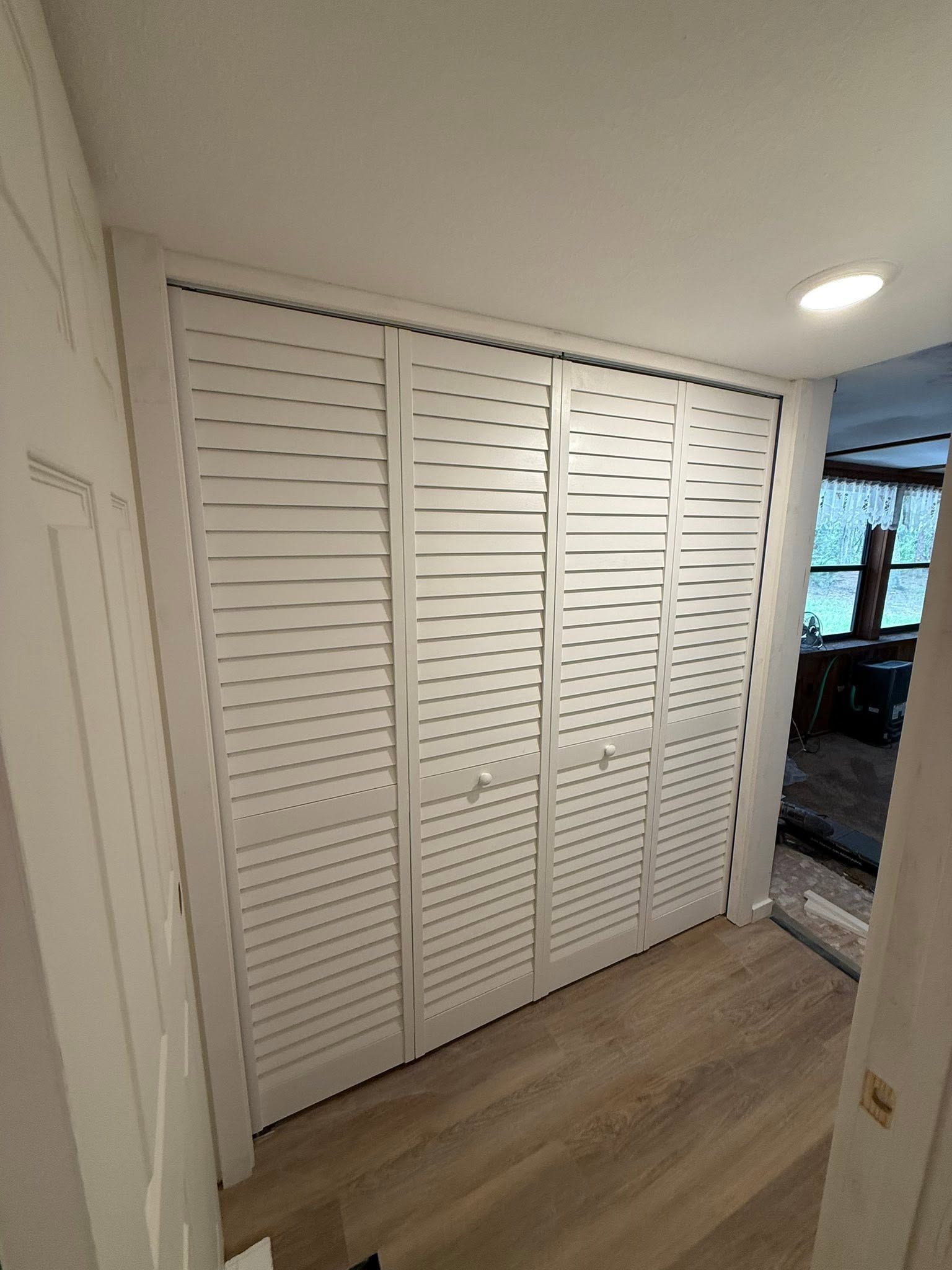 White louvered closet doors in a hallway with light wood flooring and a glimpse into another room.