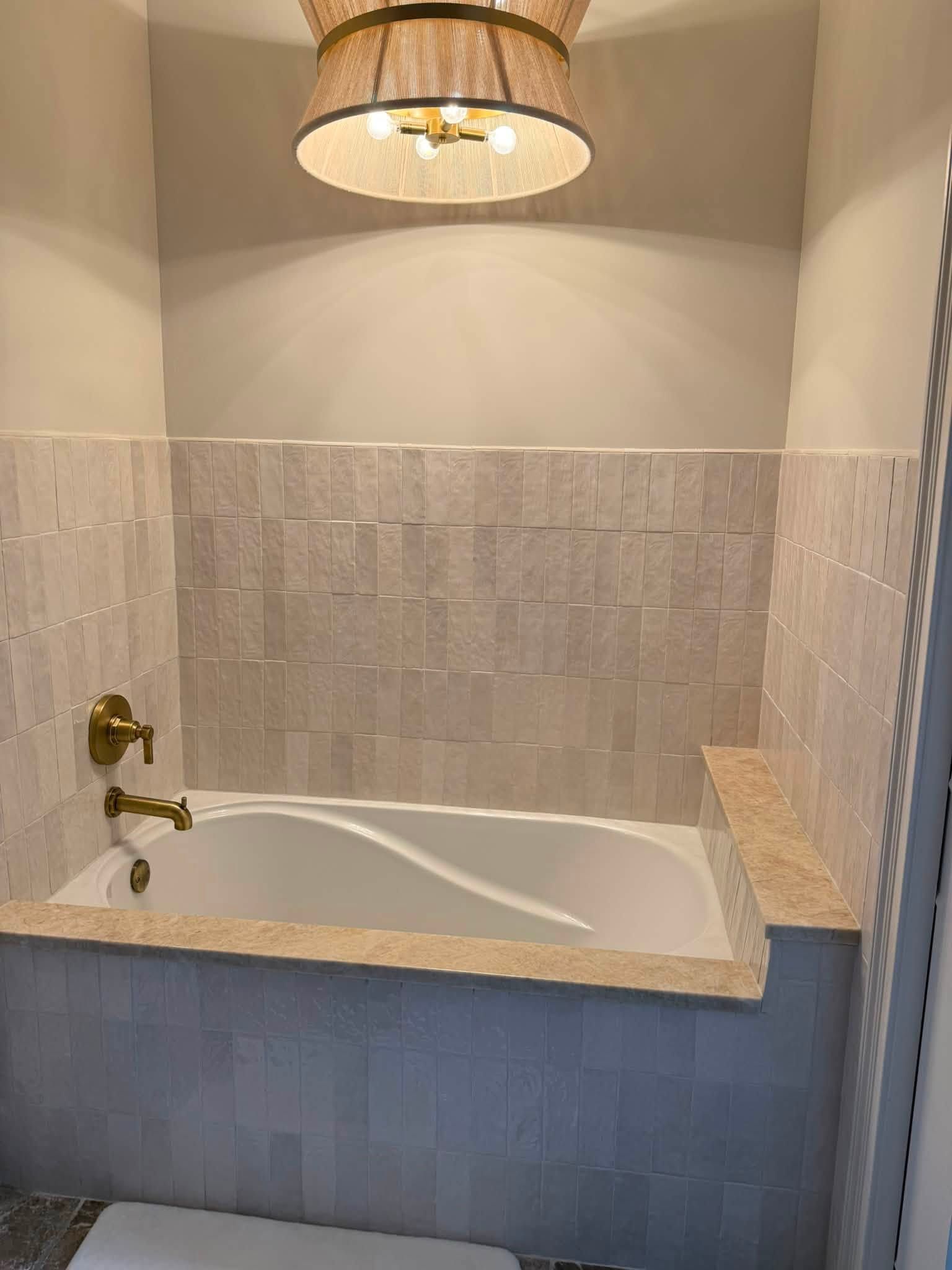 Small tiled bathroom alcove with a white bathtub, brass faucet, and woven pendant light over the tub