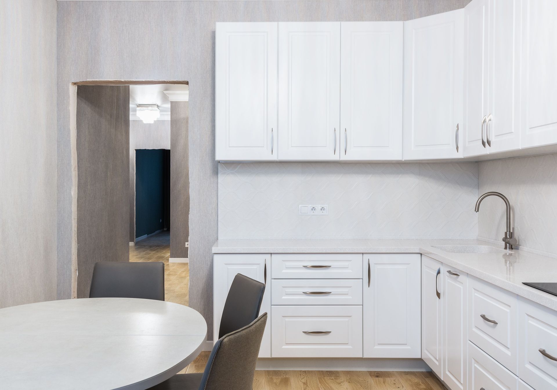 Bright white kitchen with cabinets, sink, and round table; doorway opens to a hallway.