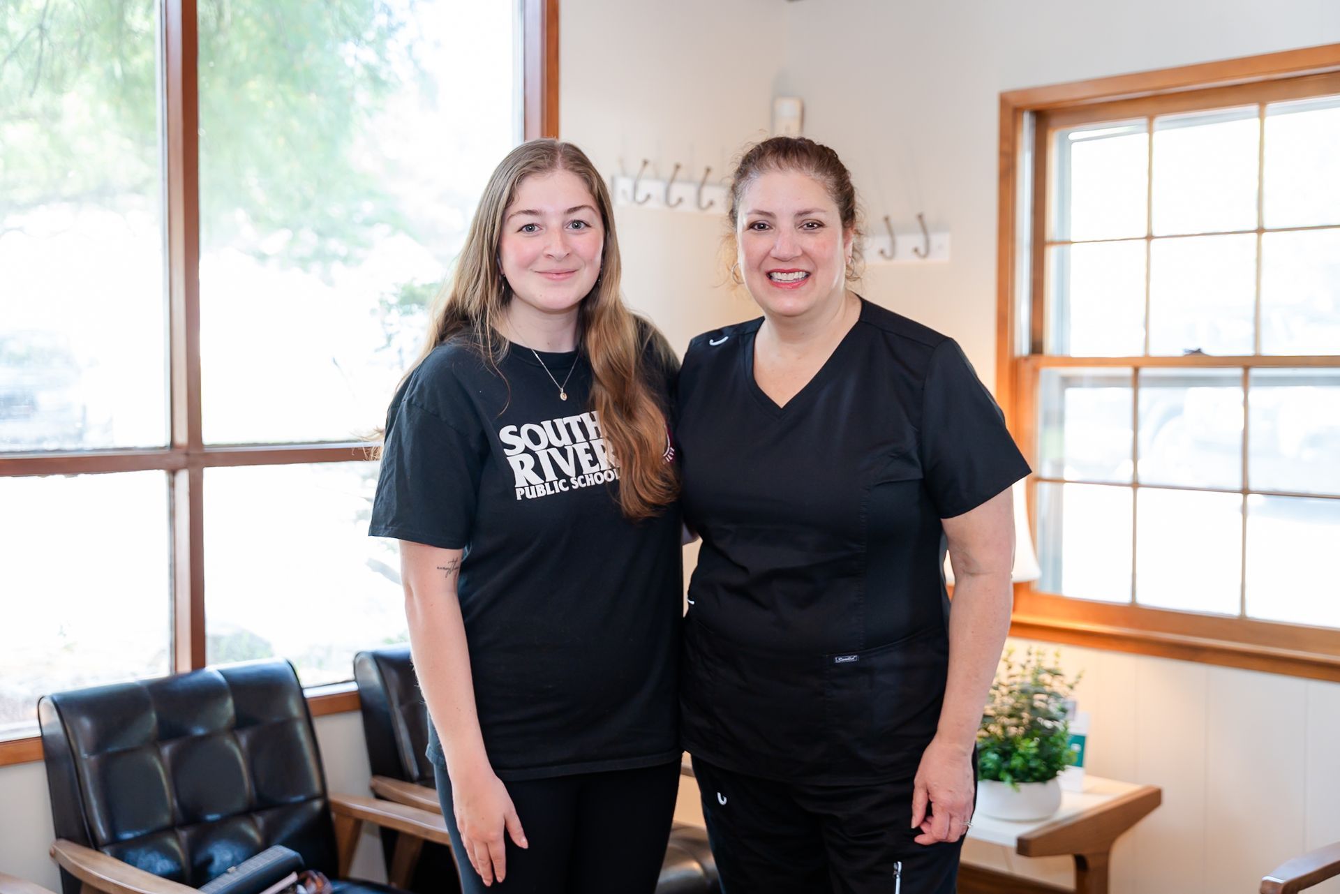 Two women in a room, smiling, one wearing a t-shirt, the other in black scrubs, next to a window.