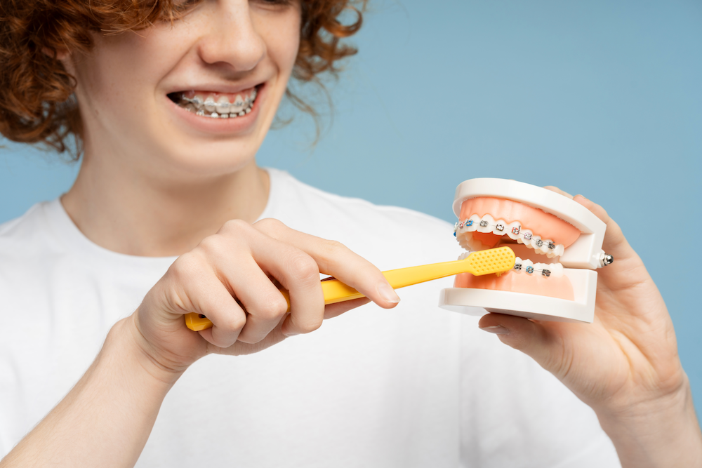 A young man is brushing his teeth with braces while holding a model of teeth.