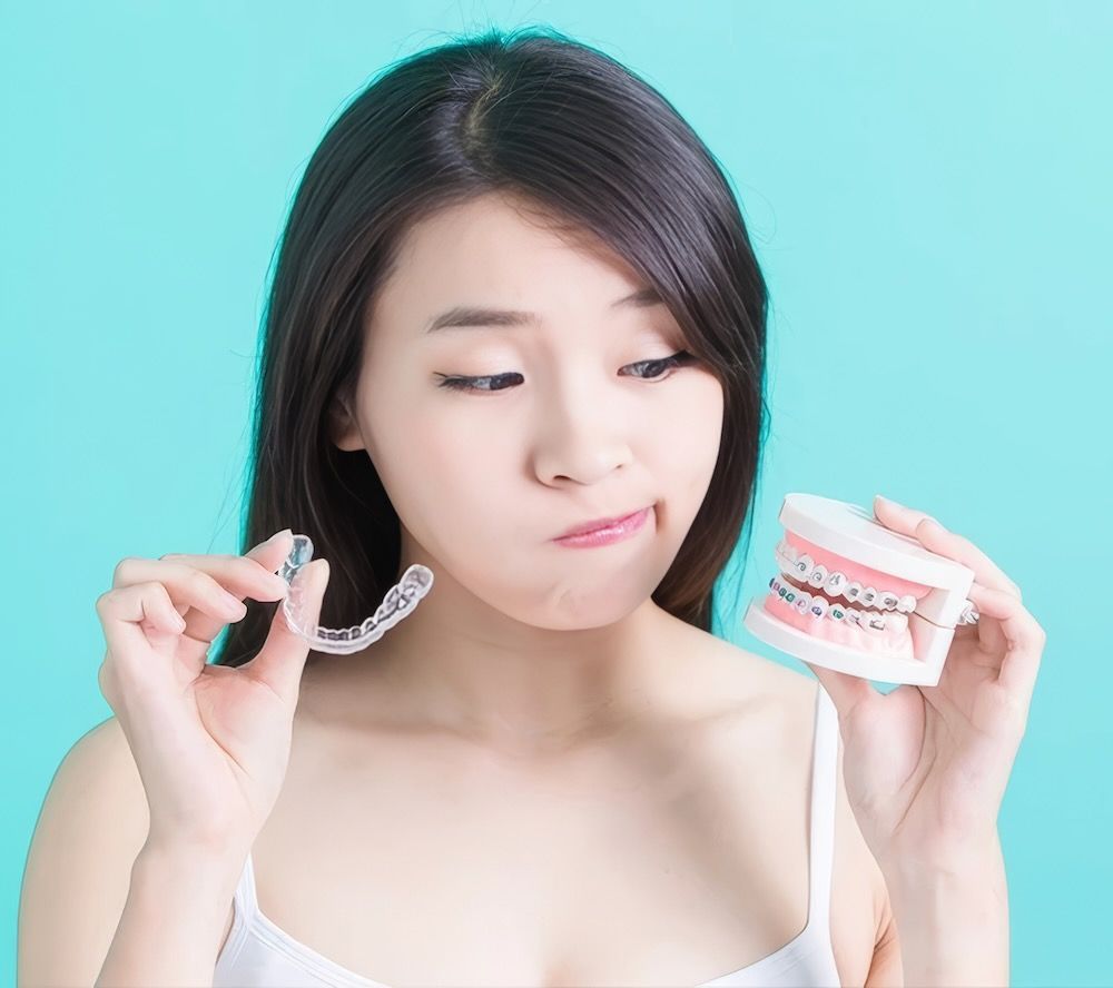 A person holds a clear dental aligner in one hand and a model of teeth with metal braces in the other against a blue wall.