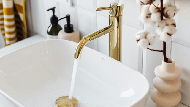 Gold faucet with running water into a white, rectangular sink. Decorative cotton branches in a vase.