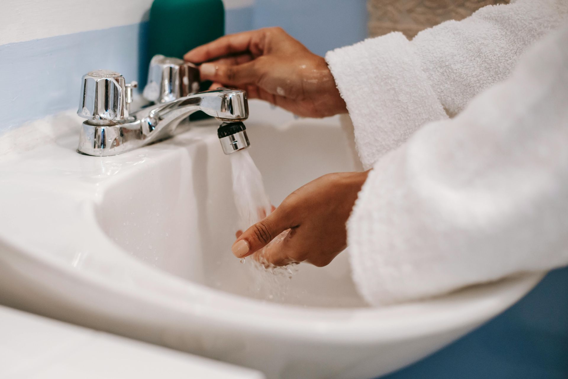 Person washing hands in a white sink under running water; wearing a white robe.