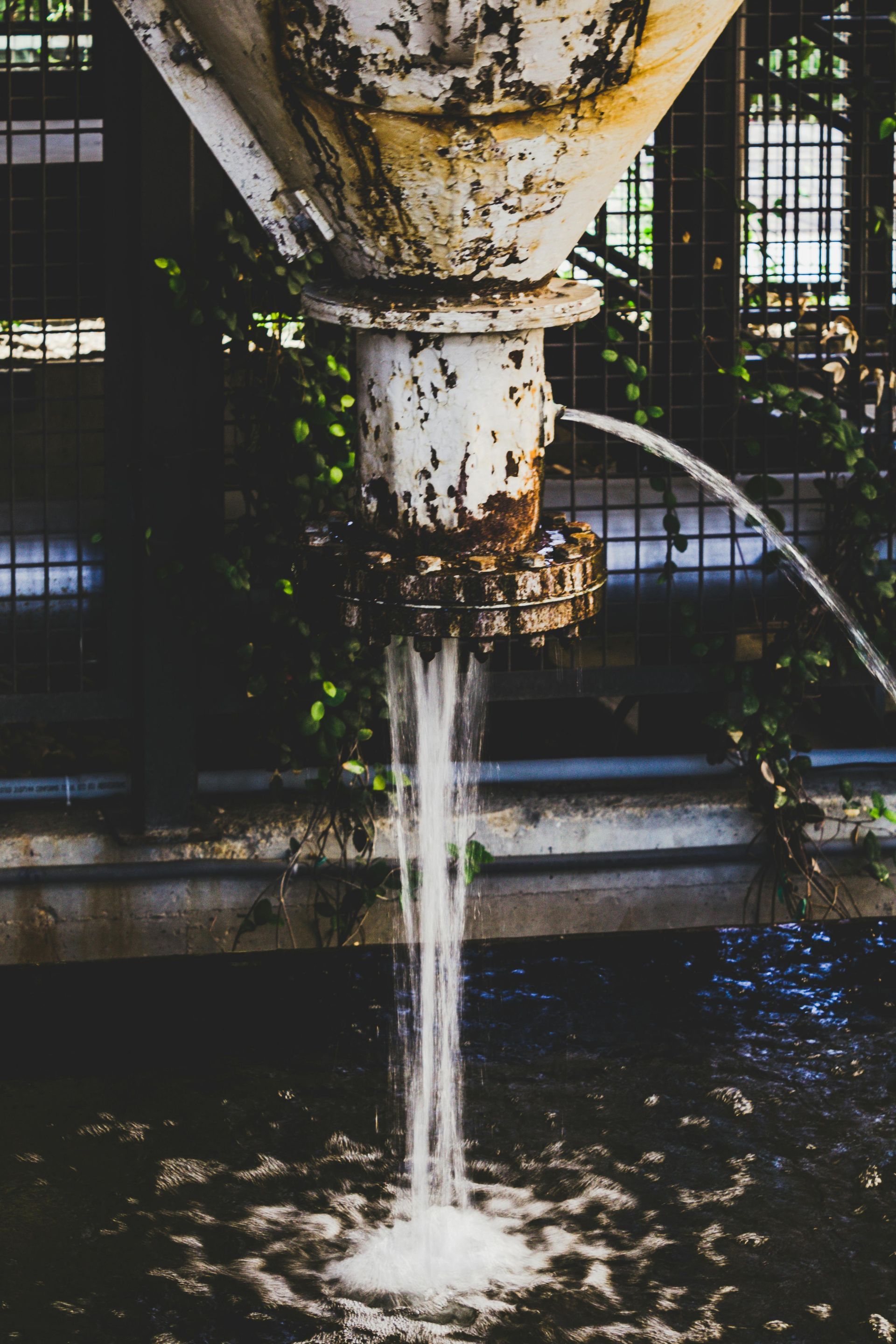 Corroded metal pipe pouring water into a dark, foamy pool outdoors.