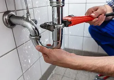 Plumber fixing a leaking sink pipe with a wrench, water spraying, near white tile.