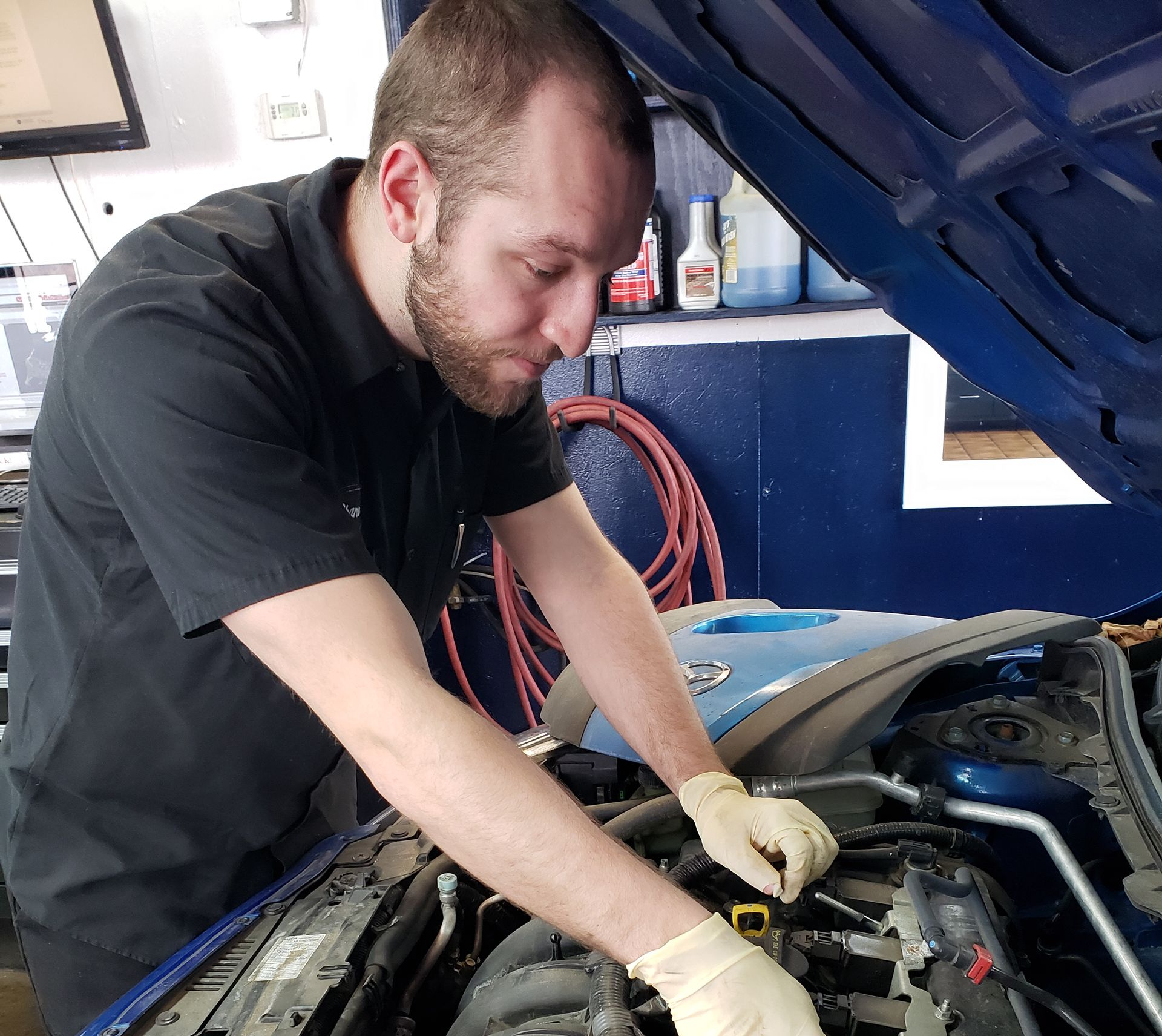 A mechanic wearing yellow gloves inspects the engine of a blue car inside a repair shop.