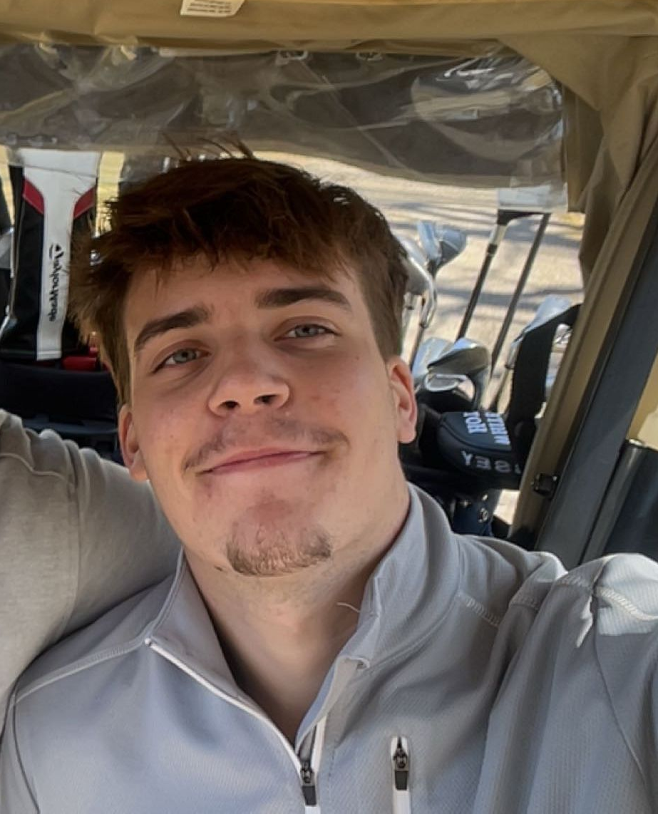 A person with short brown hair and a light beard smiles in a selfie taken inside a golf cart with clubs in the background.