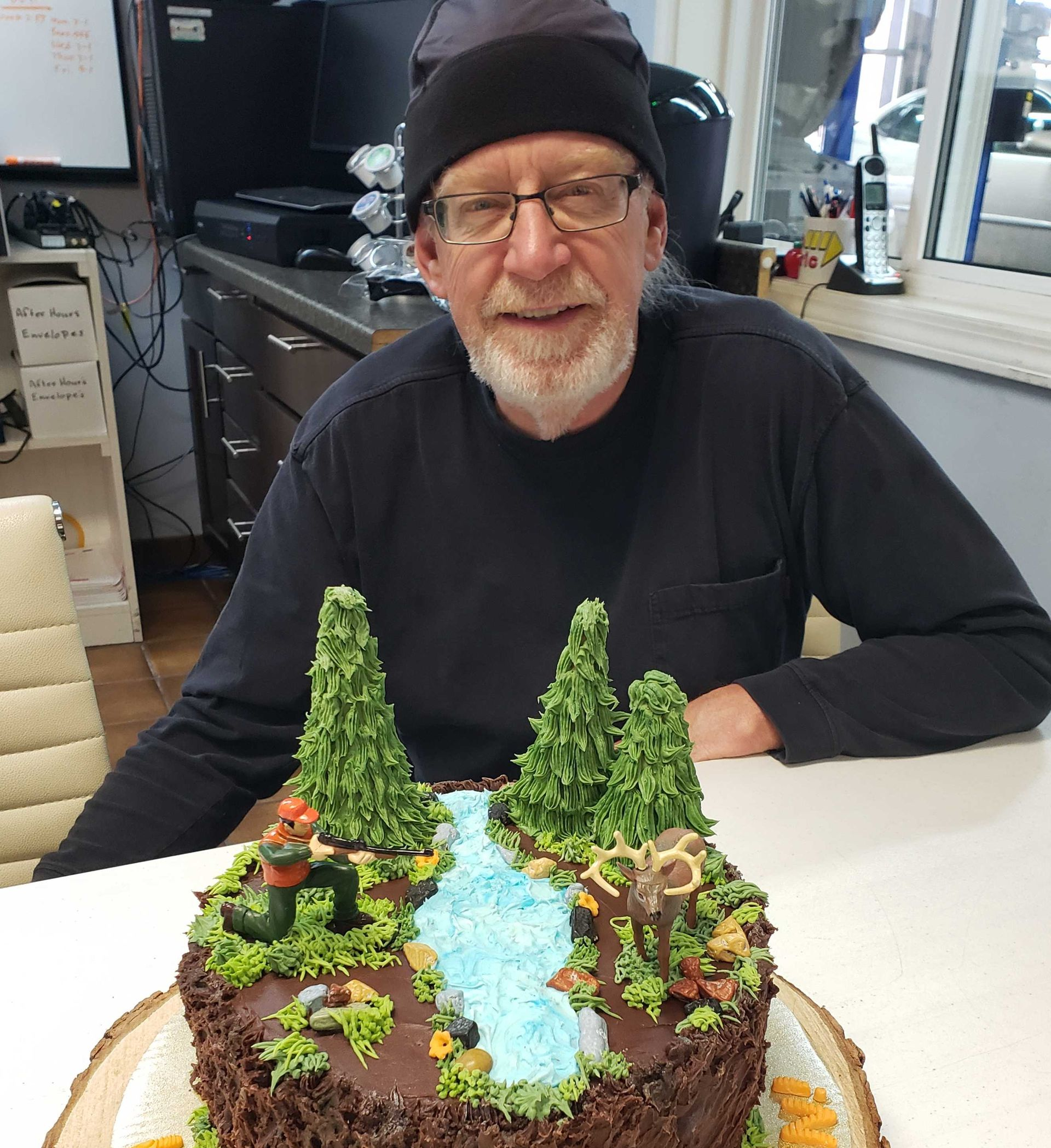 A person with a beard and dark beanie sits behind a nature-themed cake with icing trees, a river, and a small deer figure.