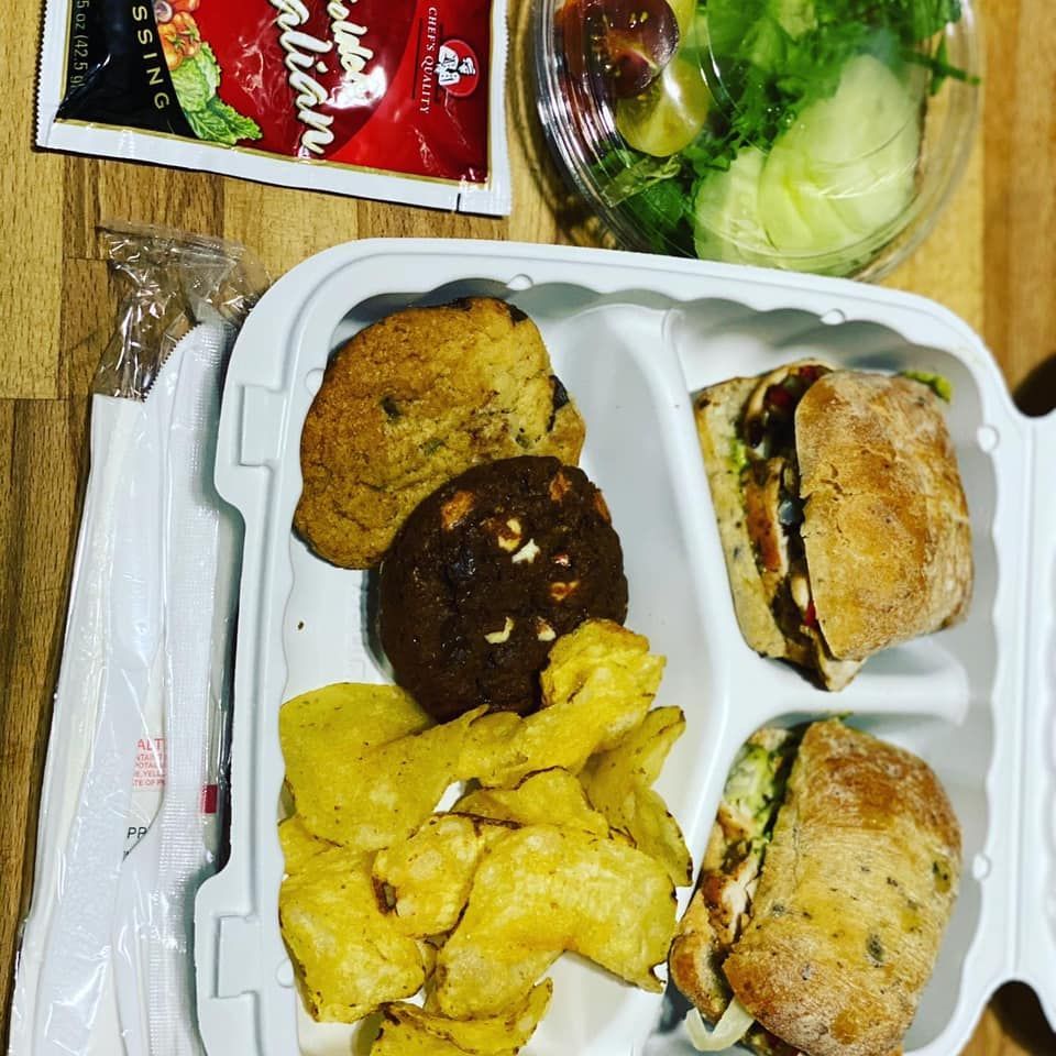 Takeout meal in a white container with fried items, sandwich halves, salad, and packaged snacks on a table
