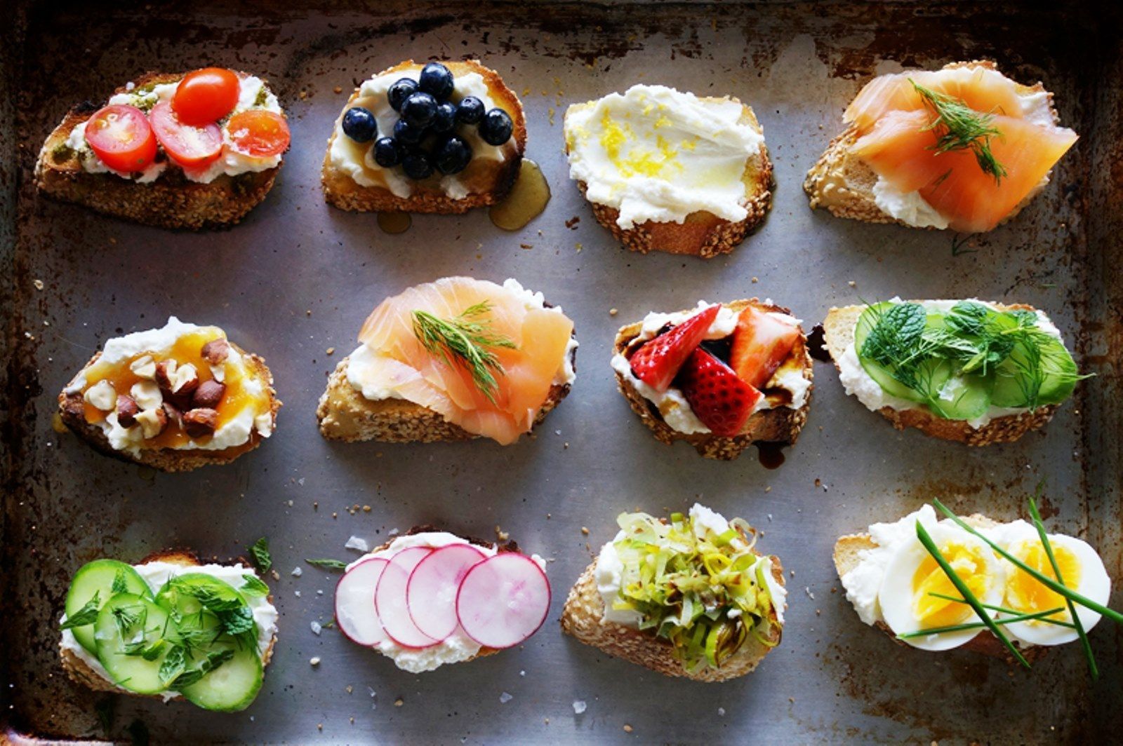 Assorted colorful open-faced sandwiches arranged on a baking tray, topped with vegetables, fish, eggs, and cheese