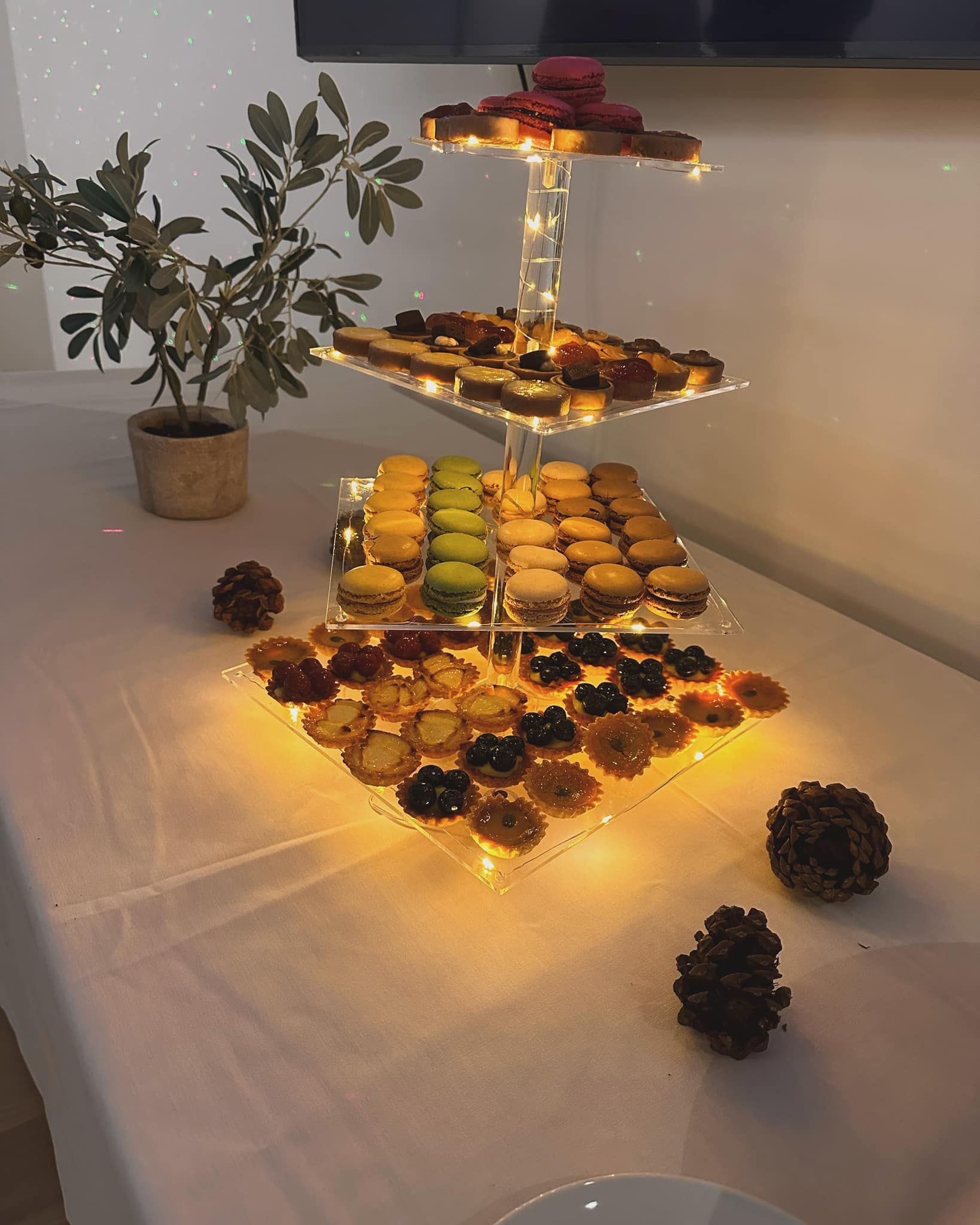 Three-tier dessert display with glowing candles and cookies on a table beside a small potted plant.
