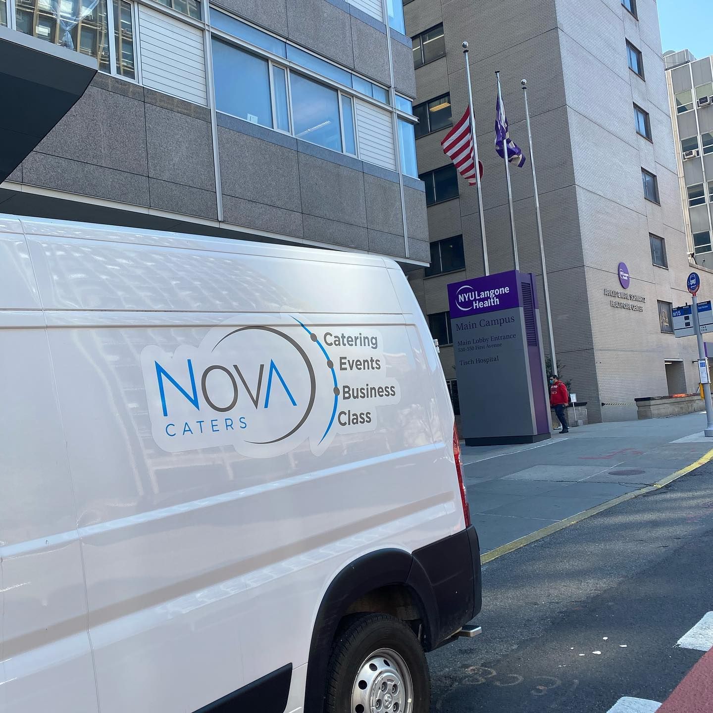 White NOVA delivery van parked on a city street beside a building with flags and a purple sign.