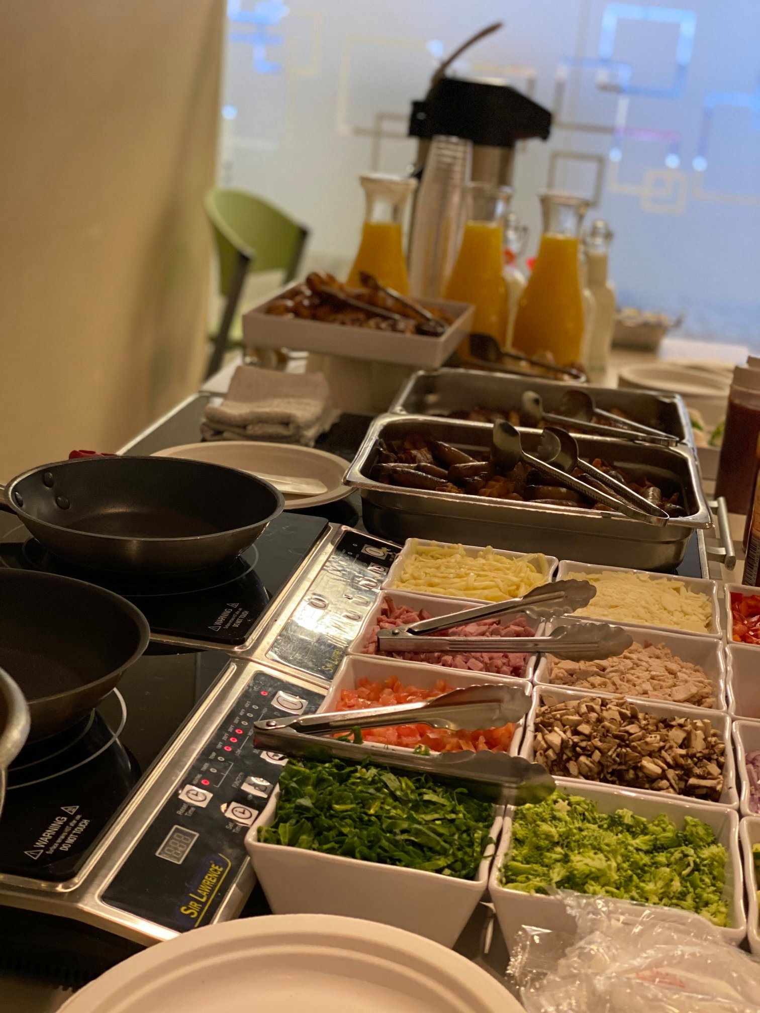 Self-serve breakfast buffet with pans, trays of toppings, and orange juice dispensers