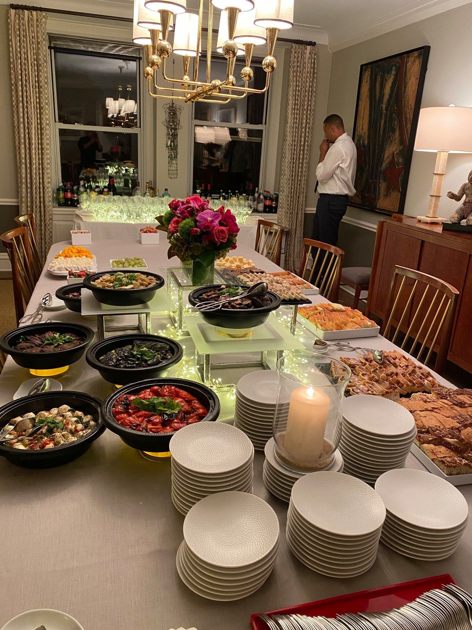 Buffet table set with bowls, plates, candles, flowers, and dishes in a dining room; a person stands in back.