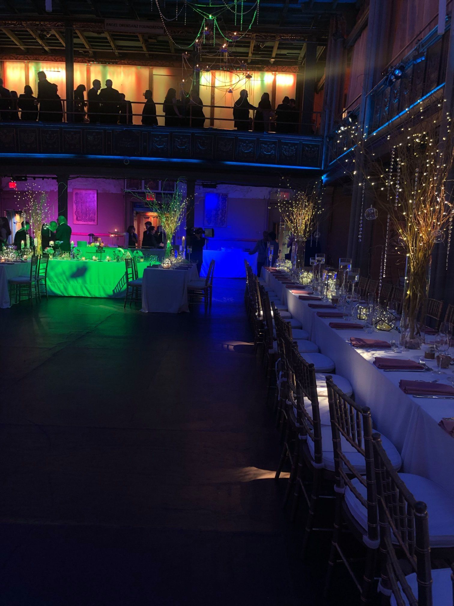 Dimly lit banquet hall with colorful stage lighting and long set tables along the right side.