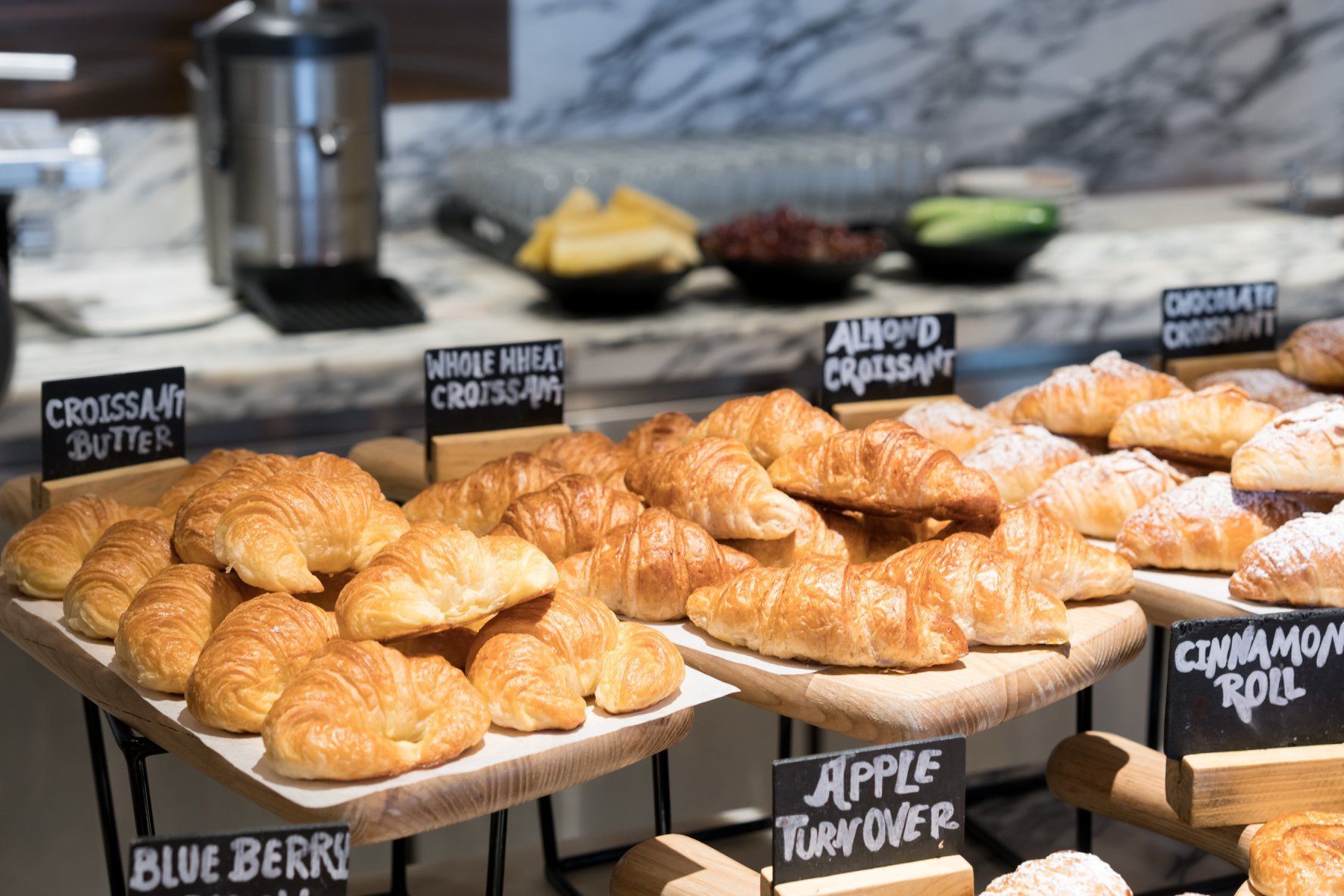 Tray of assorted pastries with label signs, including croissants and blueberry, apple turnover, and cinnamon roll pastries.
