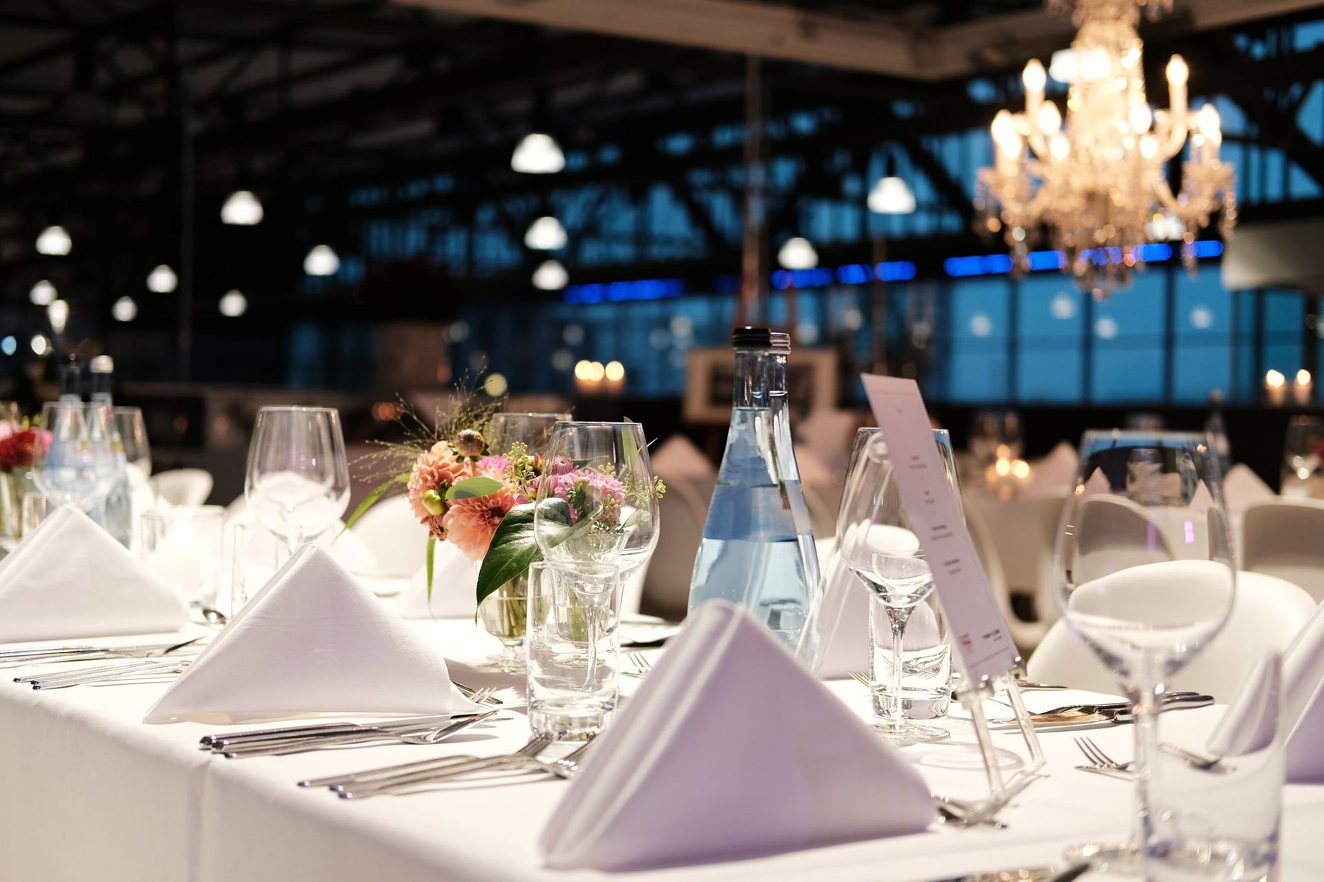 Elegant banquet table with white folded napkins, glassware, floral centerpieces, and chandeliers in a dimly lit hall