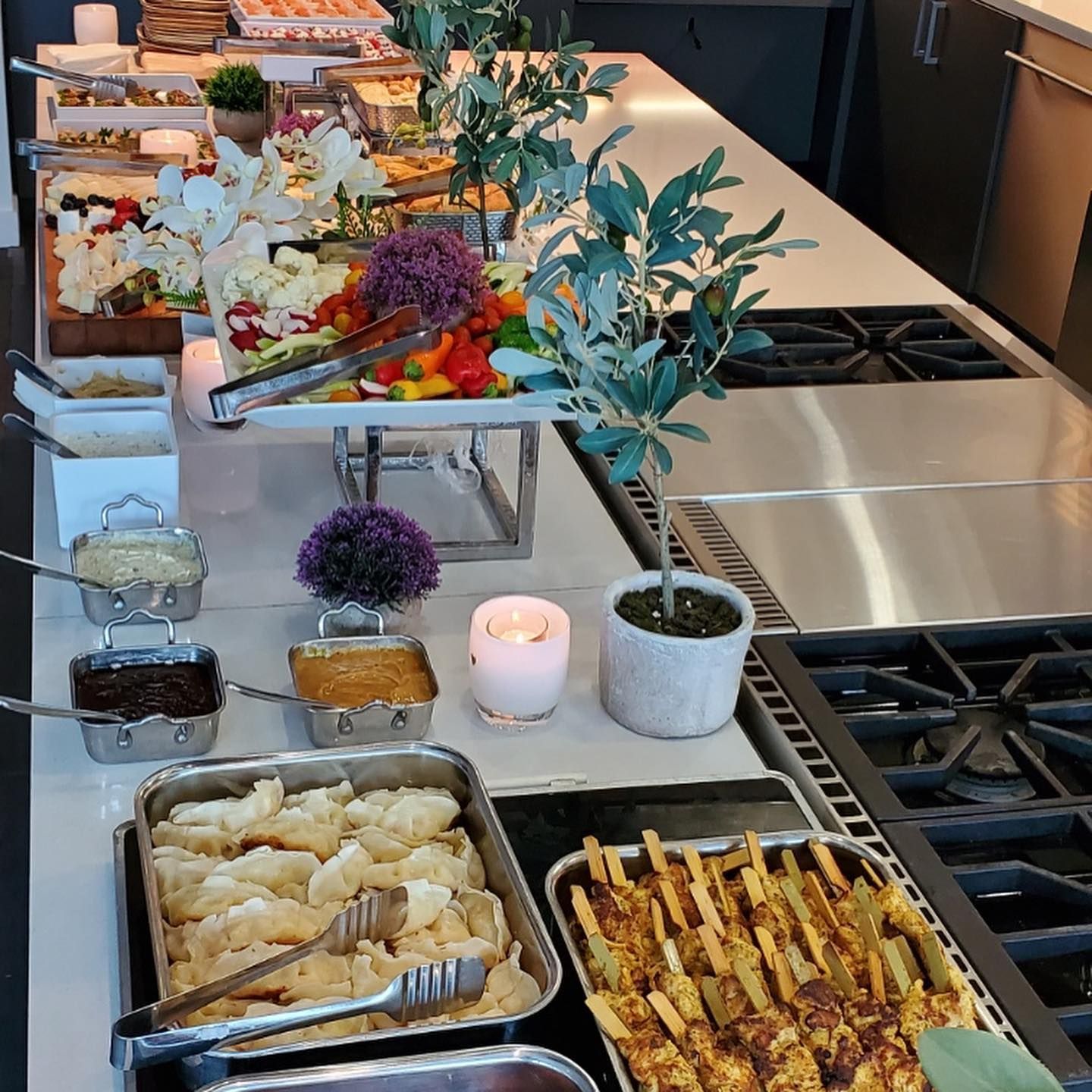 Buffet counter with fruit, cheeses, breadsticks, dips, candles, and potted decor in a modern kitchen.