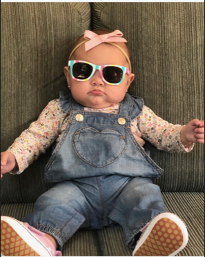 Baby in sunglasses, pink bow, and denim overalls sitting on a striped couch.